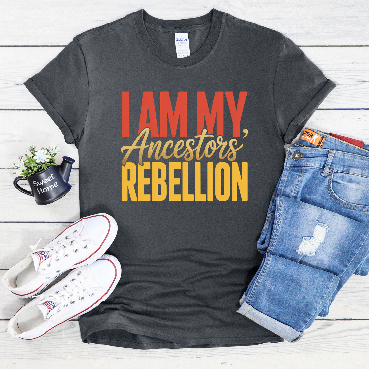 Gray t-shirt with 'I AM MY ANCESTORS REBELLION' text, white sneakers, blue jeans, and a mug on a wooden surface.