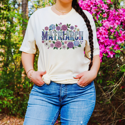 Person wearing a white t-shirt with 'Matriarch' design in a floral arrangement, standing in front of pink flowers.