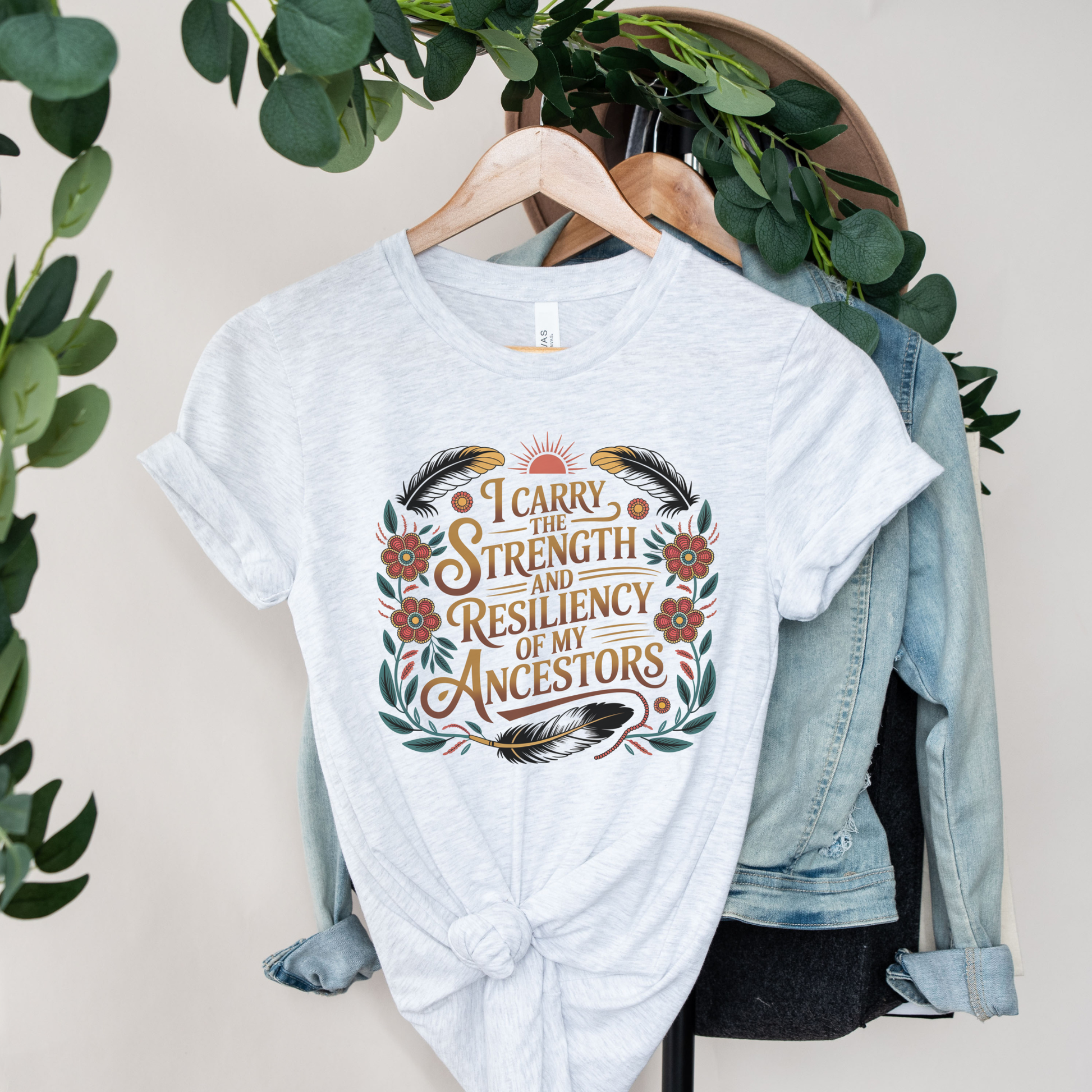  White t-shirt with motivational quote on a hanger with a denim jacket and greenery. indigenous shirt, ancestor strength shirt, resiliency shirt, indigenous inspired apparel, meaningful t shirts, strength quote shirt, heritage shirt, empowerment shirt, cultural awareness clothing, statement t shirt, social justice apparel, unity shirt, inspirational shirt, ally shirt, conscious fashion