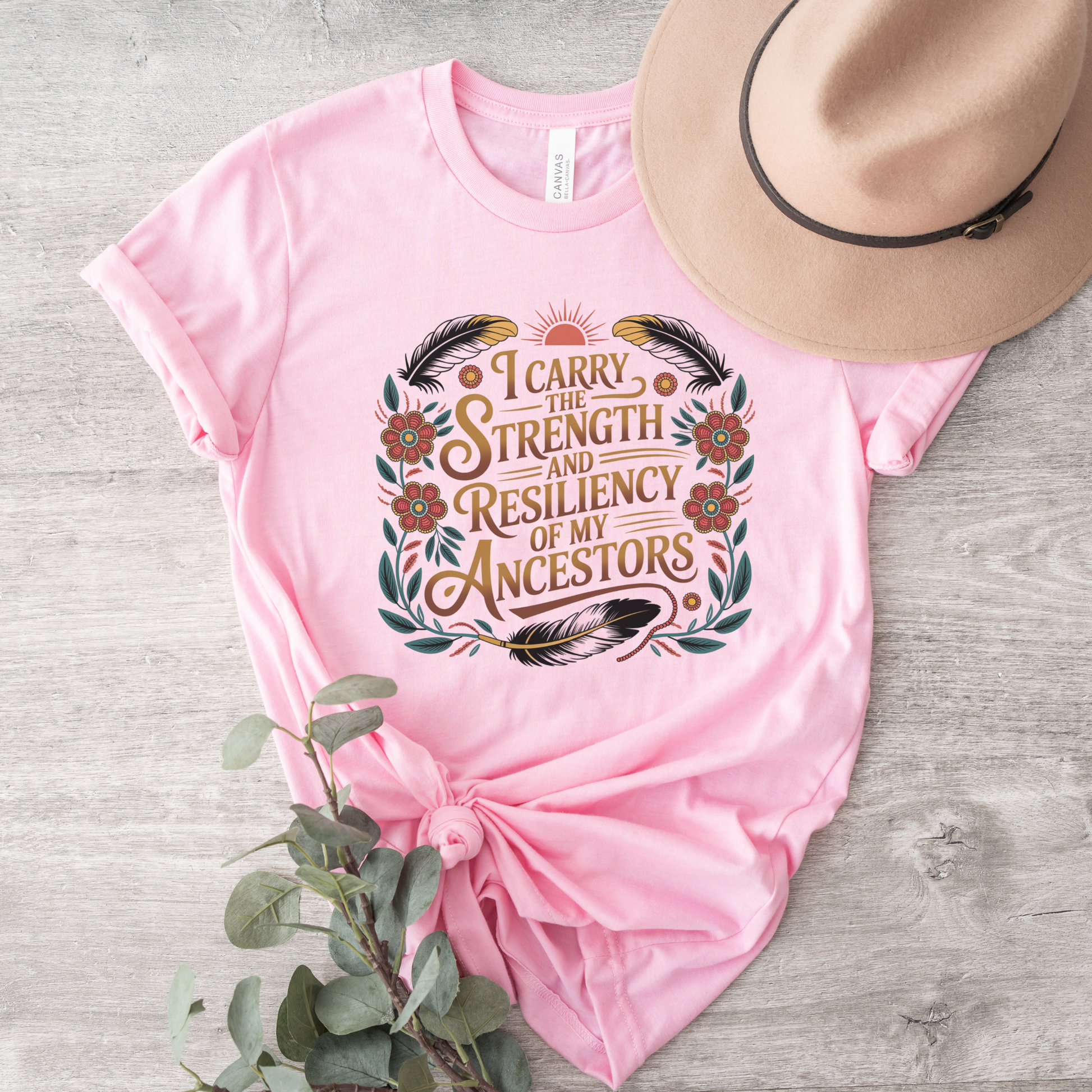 Pink t-shirt with motivational quote, hat, and plant on a wooden surface indigenous shirt, ancestor strength shirt, resiliency shirt, indigenous inspired apparel, meaningful t shirts, strength quote shirt, heritage shirt, empowerment shirt, cultural awareness clothing, statement t shirt, social justice apparel, unity shirt, inspirational shirt, ally shirt, conscious fashion