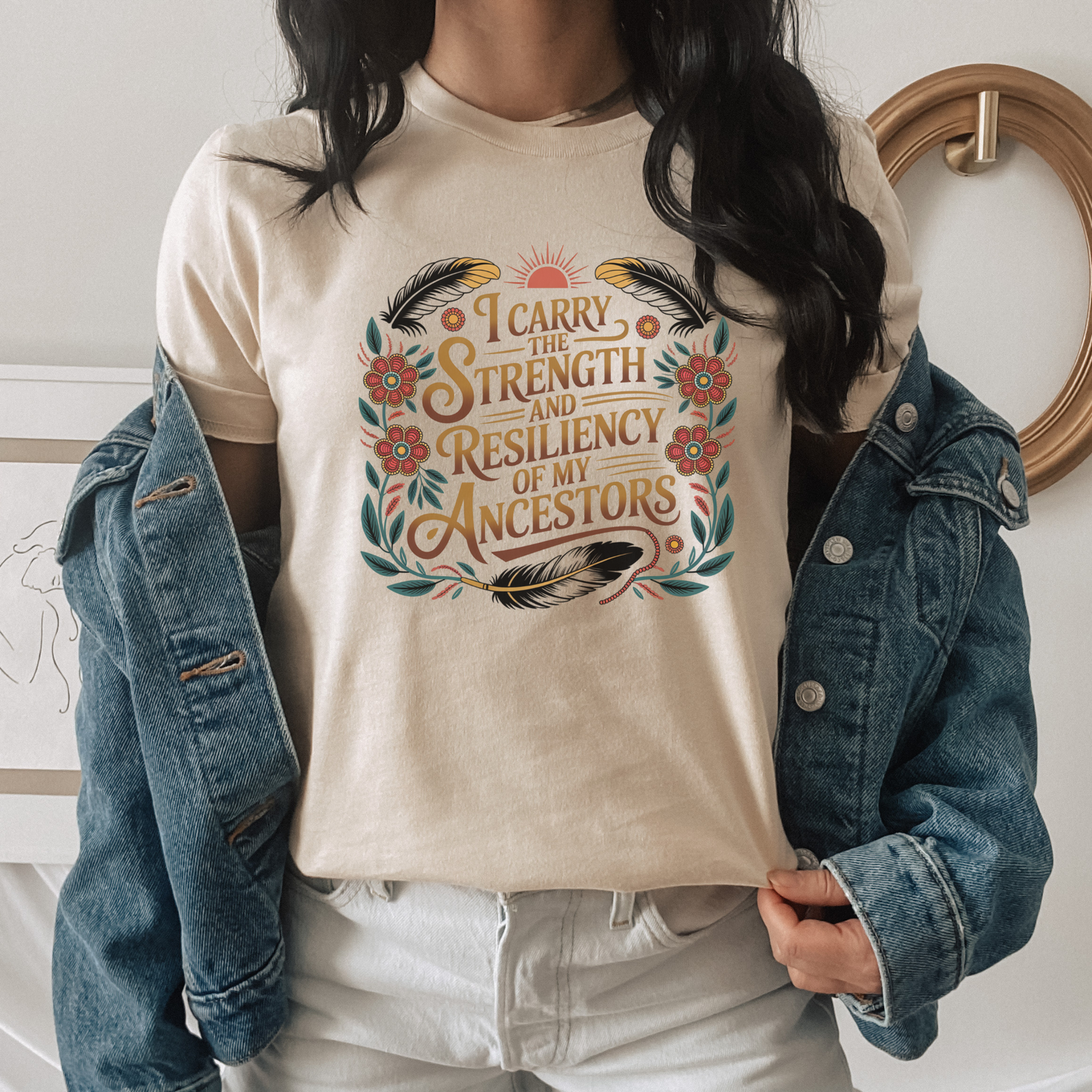 Person wearing a beige t-shirt with a motivational quote and floral design, paired with a denim jacket. indigenous shirt, ancestor strength shirt, resiliency shirt, indigenous inspired apparel, meaningful t shirts, strength quote shirt, heritage shirt, empowerment shirt, cultural awareness clothing, statement t shirt, social justice apparel, unity shirt, inspirational shirt, ally shirt, conscious fashion