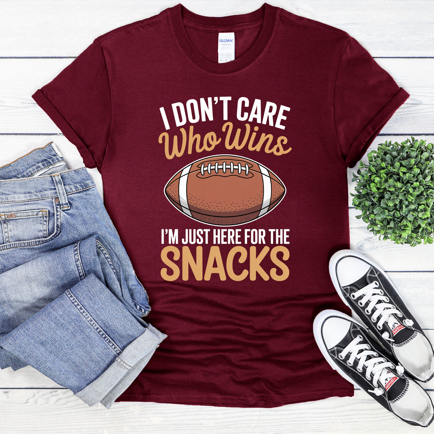Maroon t-shirt with football graphic and text, paired with jeans and sneakers on a white wooden surface. funny football shirt, game day shirt, snack lover shirt, super bowl party tee, I don’t care who wins shirt, here for the snacks shirt, football fan t-shirt, tailgate outfit, big game shirt, casual sports fan tee, humorous graphic tee, unisex game day top, football party clothing, lazy fan shirt, game day snacks shirt