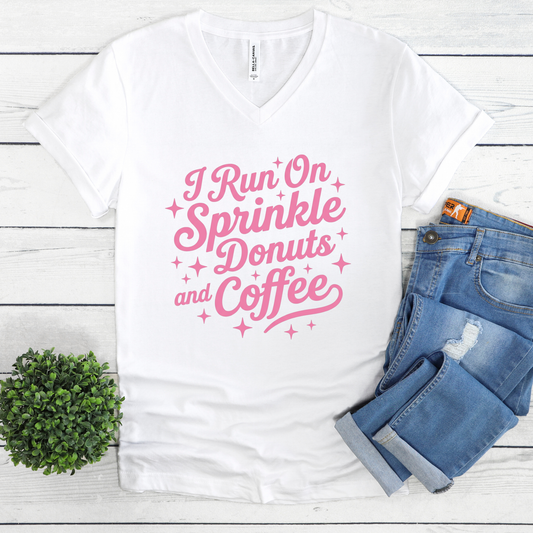 White t-shirt with pink text 'I Run On Sprinkle Donuts and Coffee' on a wooden surface with jeans and a plant.
