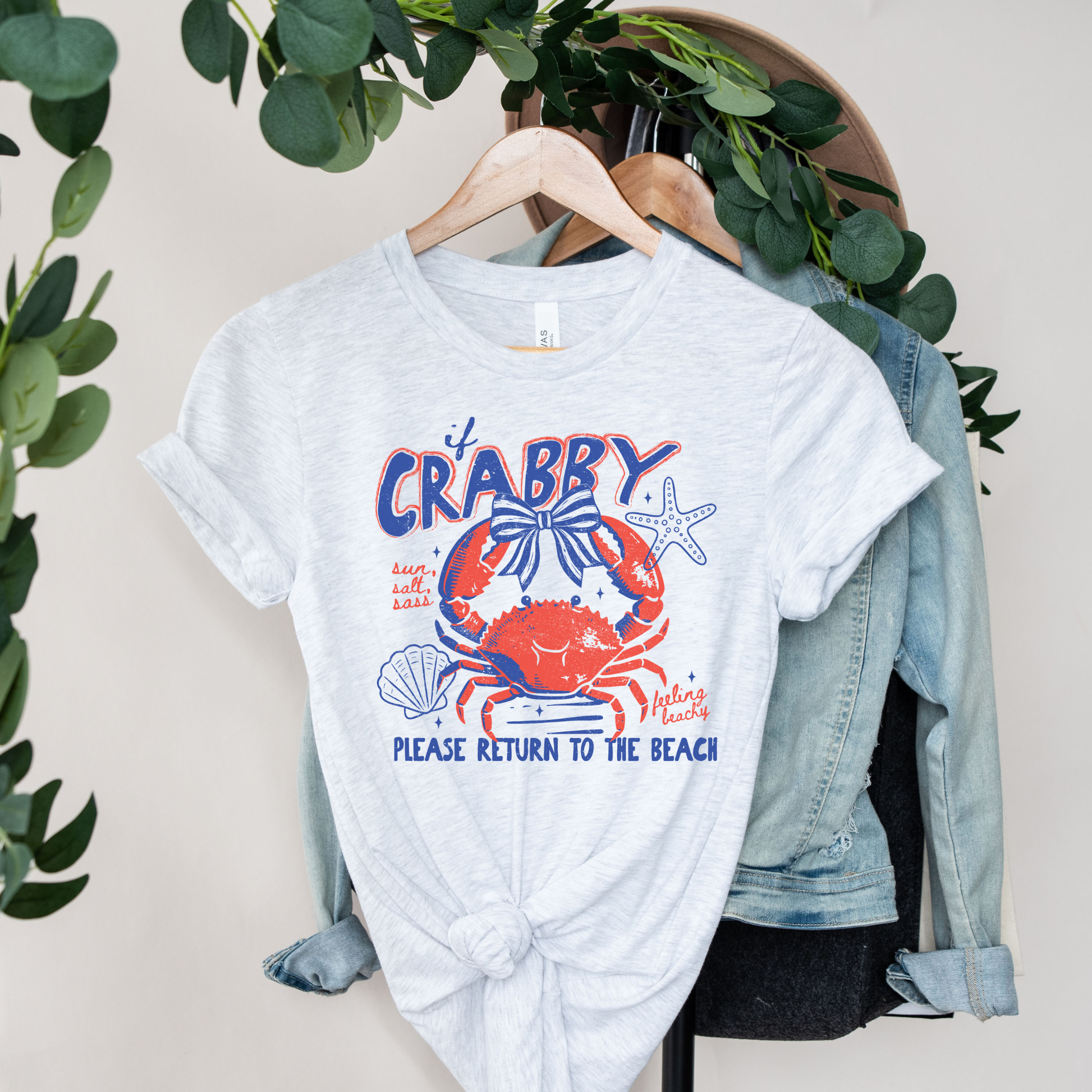 White t-shirt with a colorful crab graphic and text on a hanger with a denim jacket and greenery. beach shirt, coastal tee, funny beach shirt, beach vacation outfit, summer shirt, ocean lover gift, seaside apparel, crab shirt, vacation t-shirt, casual beachwear, coastal clothing, lake life shirt, beach humor tee, seaside gifts, unisex beach shirt