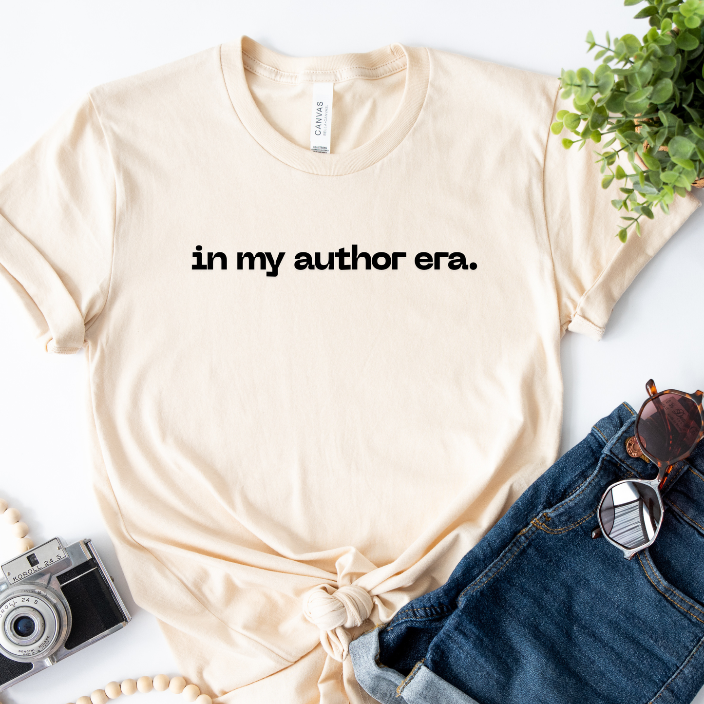 Beige t-shirt with 'in my author era.' text, sunglasses, camera, and plant on a white surface. in my author era shirt, writer shirt, author tee, writing shirt, nanowrimo shirt, book author apparel, novelist shirt, writer gift shirt, author era tee, creative writer clothing, book launch shirt, writing community tee, aspiring author shirt, fiction writer apparel, manuscript shirt