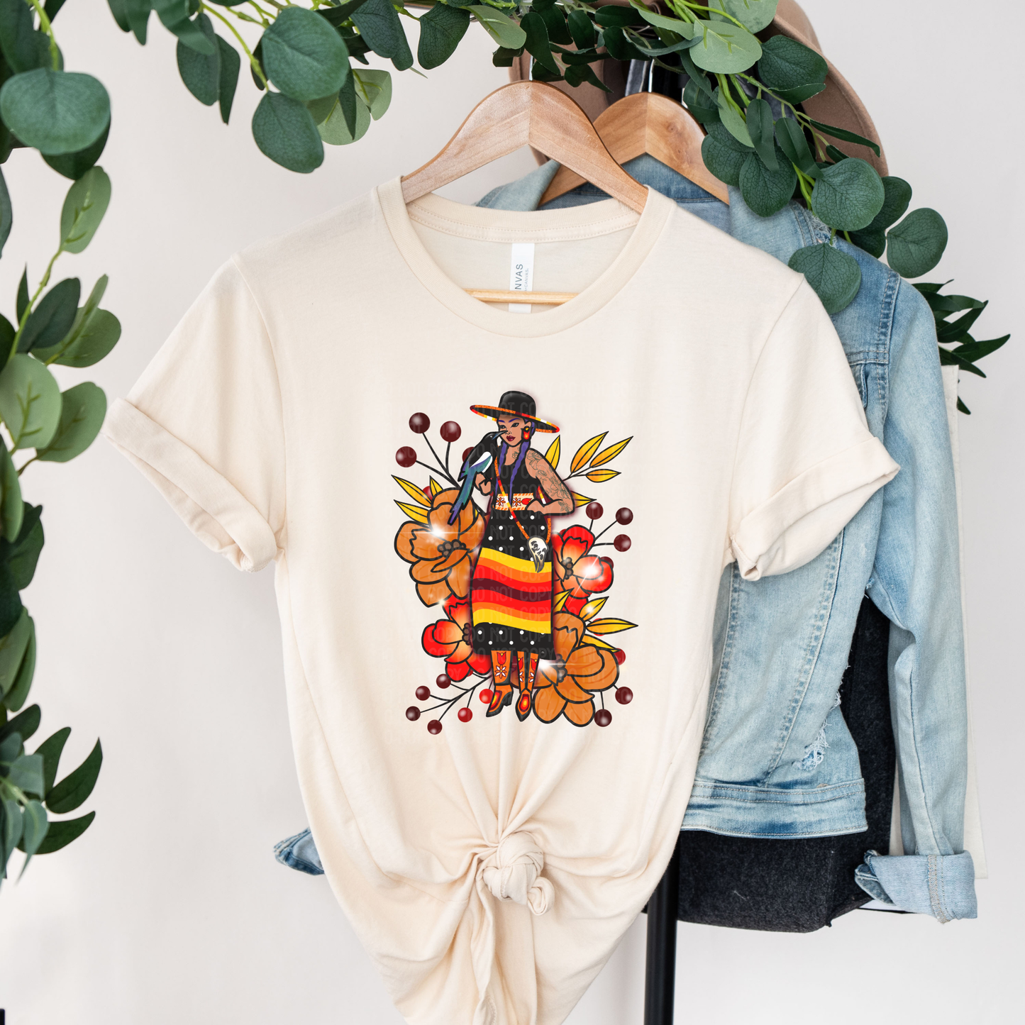 A cream-colored short-sleeve t-shirt with a graphic of a woman playing a guitar surrounded by autumn leaves, hanging on a wooden hanger next to a light blue denim jacket.