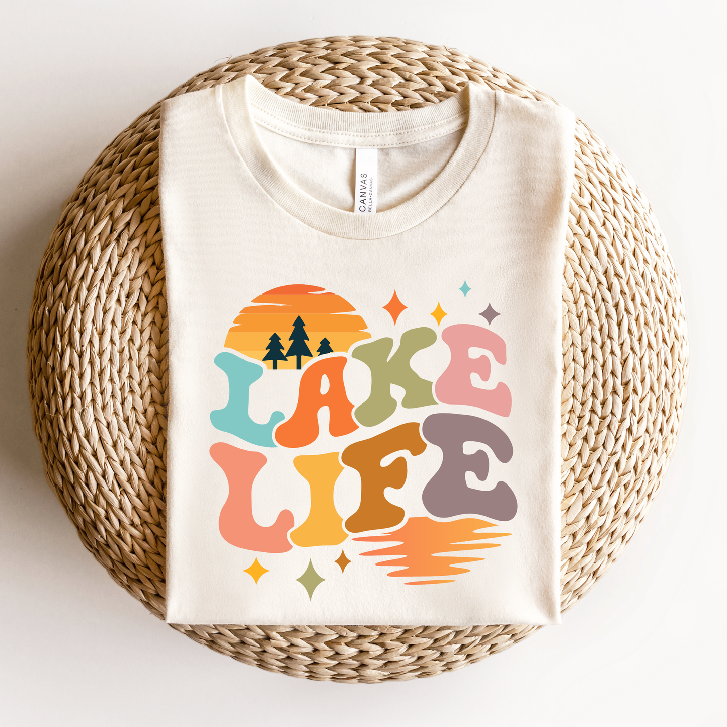 Beige t-shirt with 'Lake Life' design on a woven ottoman. lake life shirt, lake life tee, retro lake graphic shirt, sunset lake shirt, boating shirt, lake lover apparel, summer lake tee, unisex lake life clothing, lake house gift, fishing shirt, cottage lake outfit, lake vacation tee, boater shirt, lakeside apparel, retro sunset graphic tee