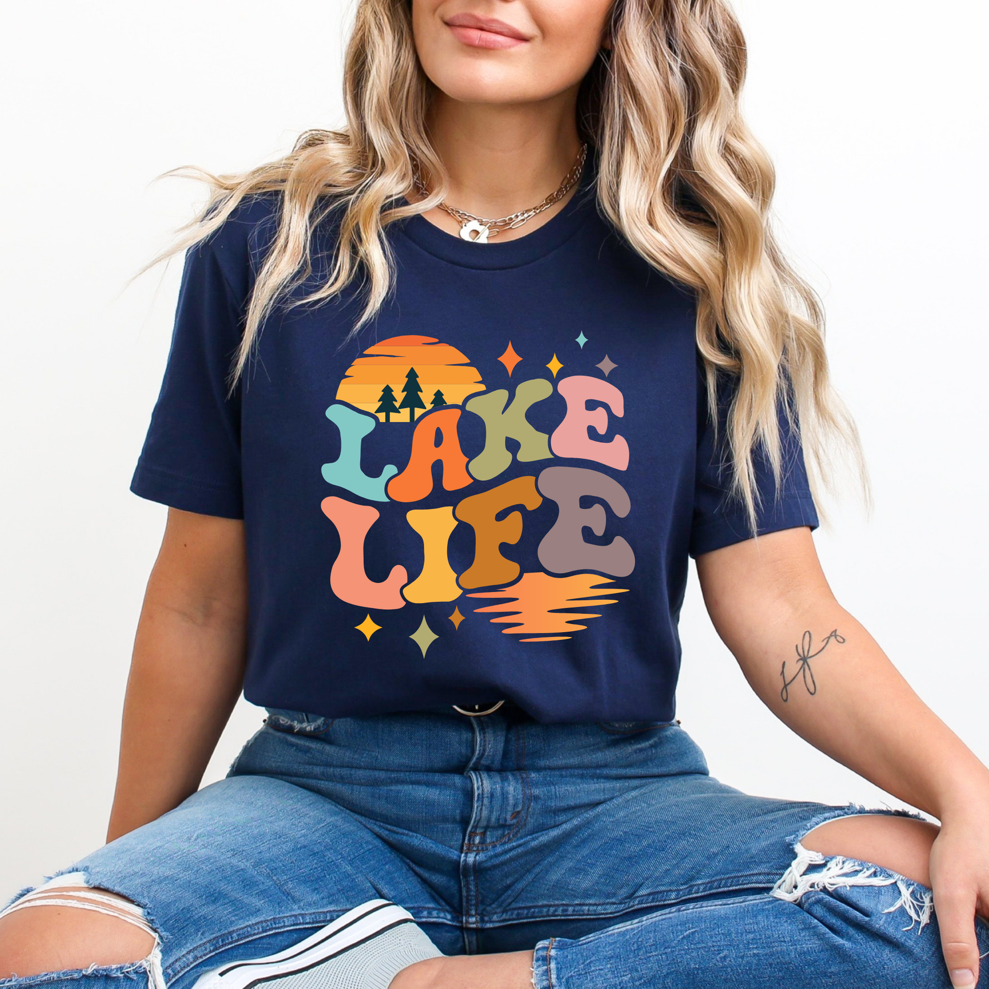 Person wearing a navy blue t-shirt with 'Lake Life' colorful design. lake life shirt, lake life tee, retro lake graphic shirt, sunset lake shirt, boating shirt, lake lover apparel, summer lake tee, unisex lake life clothing, lake house gift, fishing shirt, cottage lake outfit, lake vacation tee, boater shirt, lakeside apparel, retro sunset graphic tee