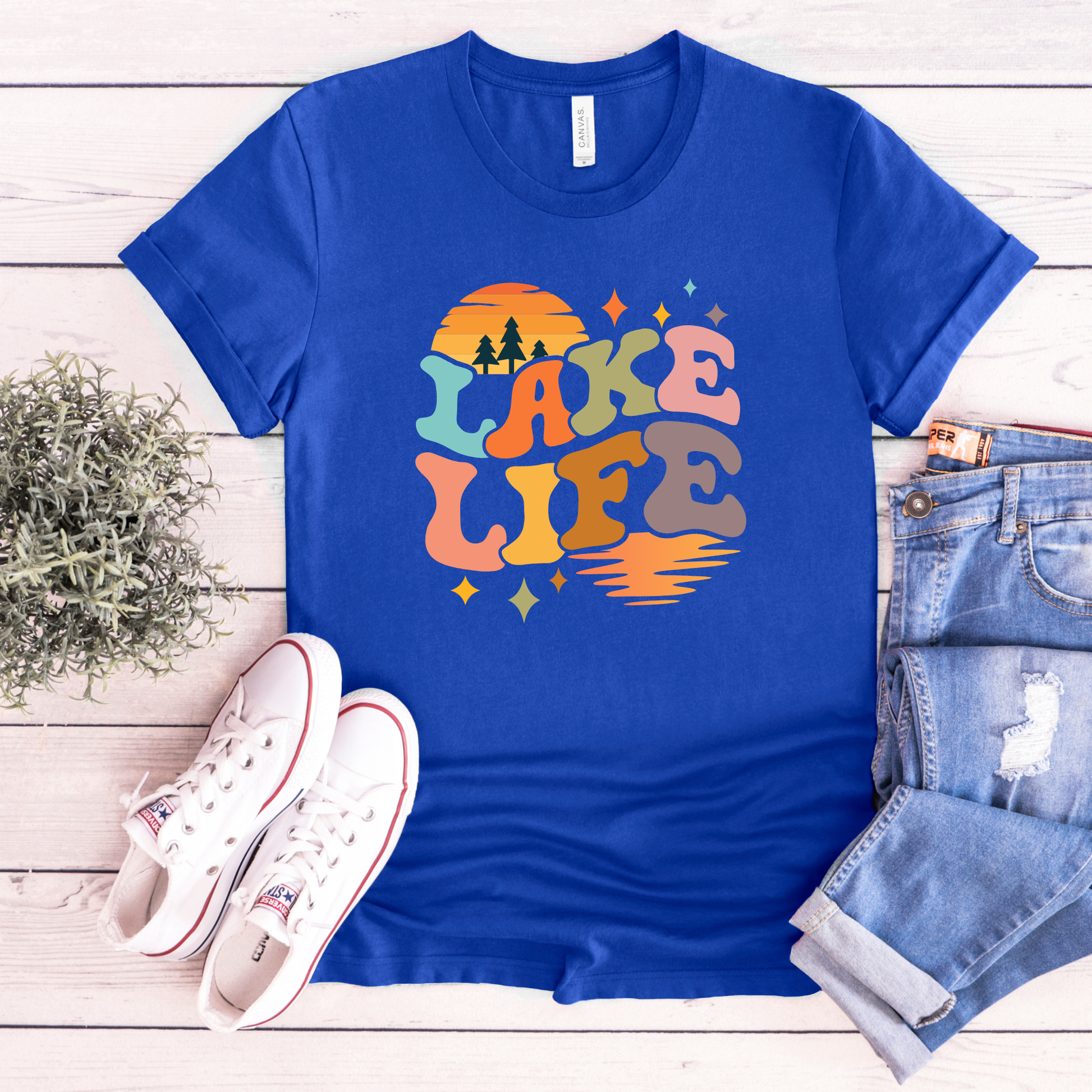 Blue t-shirt with 'Lake Life' design on a wooden surface with sneakers and jeans. lake life shirt, lake life tee, retro lake graphic shirt, sunset lake shirt, boating shirt, lake lover apparel, summer lake tee, unisex lake life clothing, lake house gift, fishing shirt, cottage lake outfit, lake vacation tee, boater shirt, lakeside apparel, retro sunset graphic tee