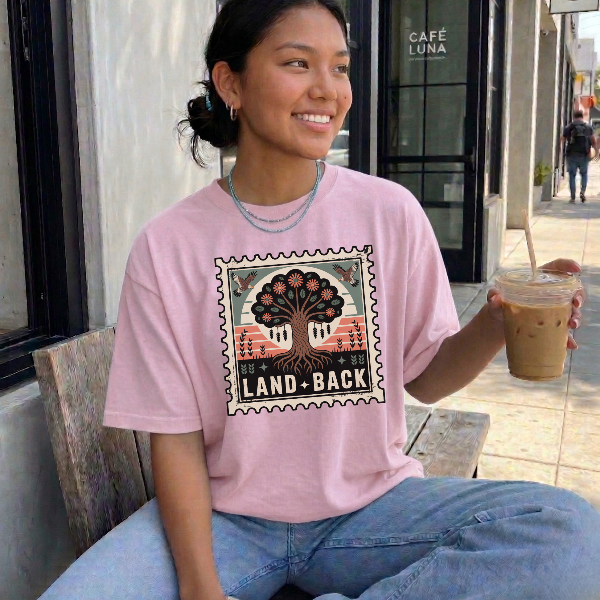 Person wearing a pink t-shirt with a graphic design, sitting outdoors holding a drink. land back shirt, indigenous awareness shirt, nature shirt, tree graphic tee, minimalist shirt, advocacy apparel, eco conscious clothing, indigenous inspired apparel, social justice shirt, unisex t shirt, earthy aesthetic clothing, statement tee