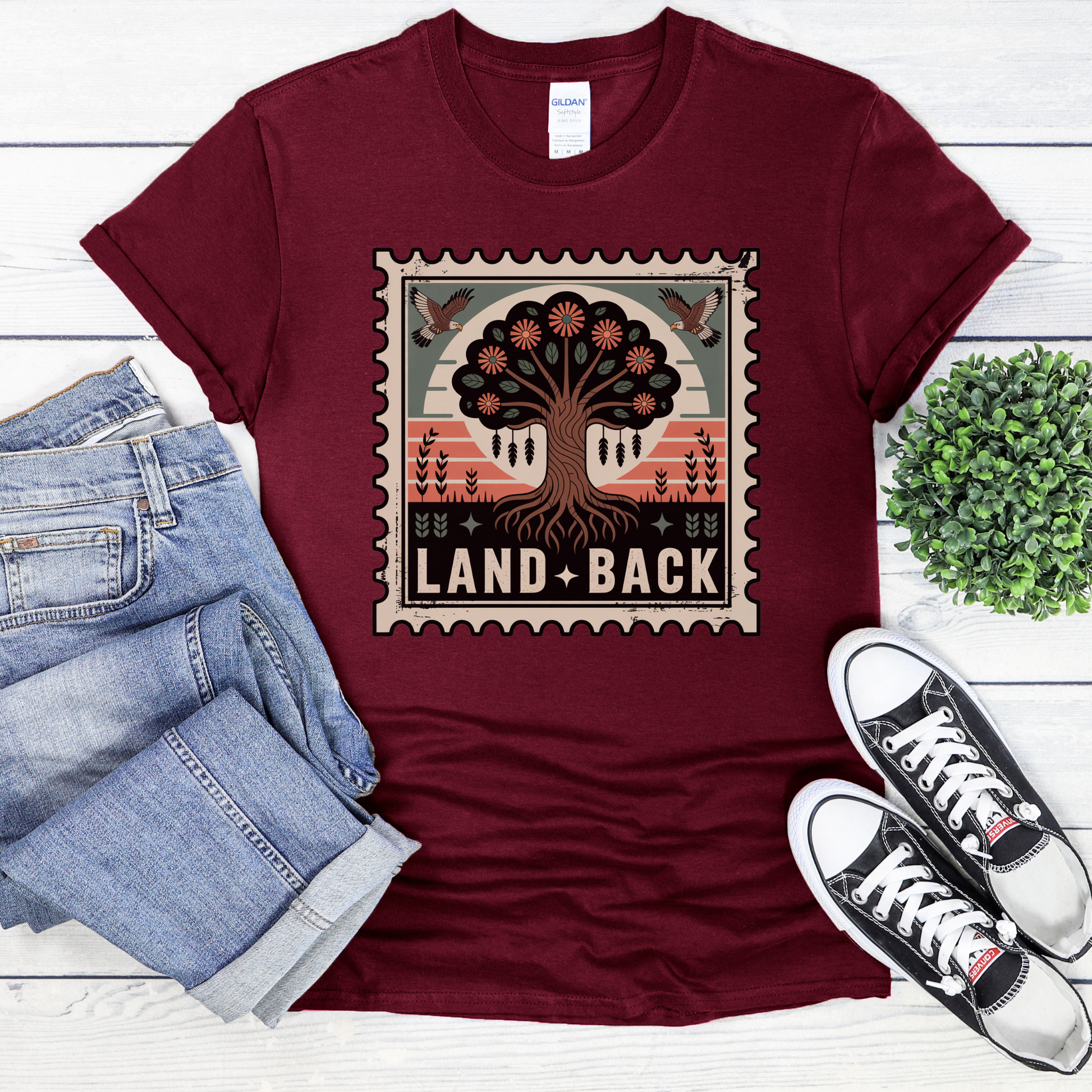 Maroon t-shirt with 'Land Back' graphic design on a white wooden surface with jeans and sneakers. land back shirt, indigenous awareness shirt, nature shirt, tree graphic tee, minimalist shirt, advocacy apparel, eco conscious clothing, indigenous inspired apparel, social justice shirt, unisex t shirt, earthy aesthetic clothing, statement tee