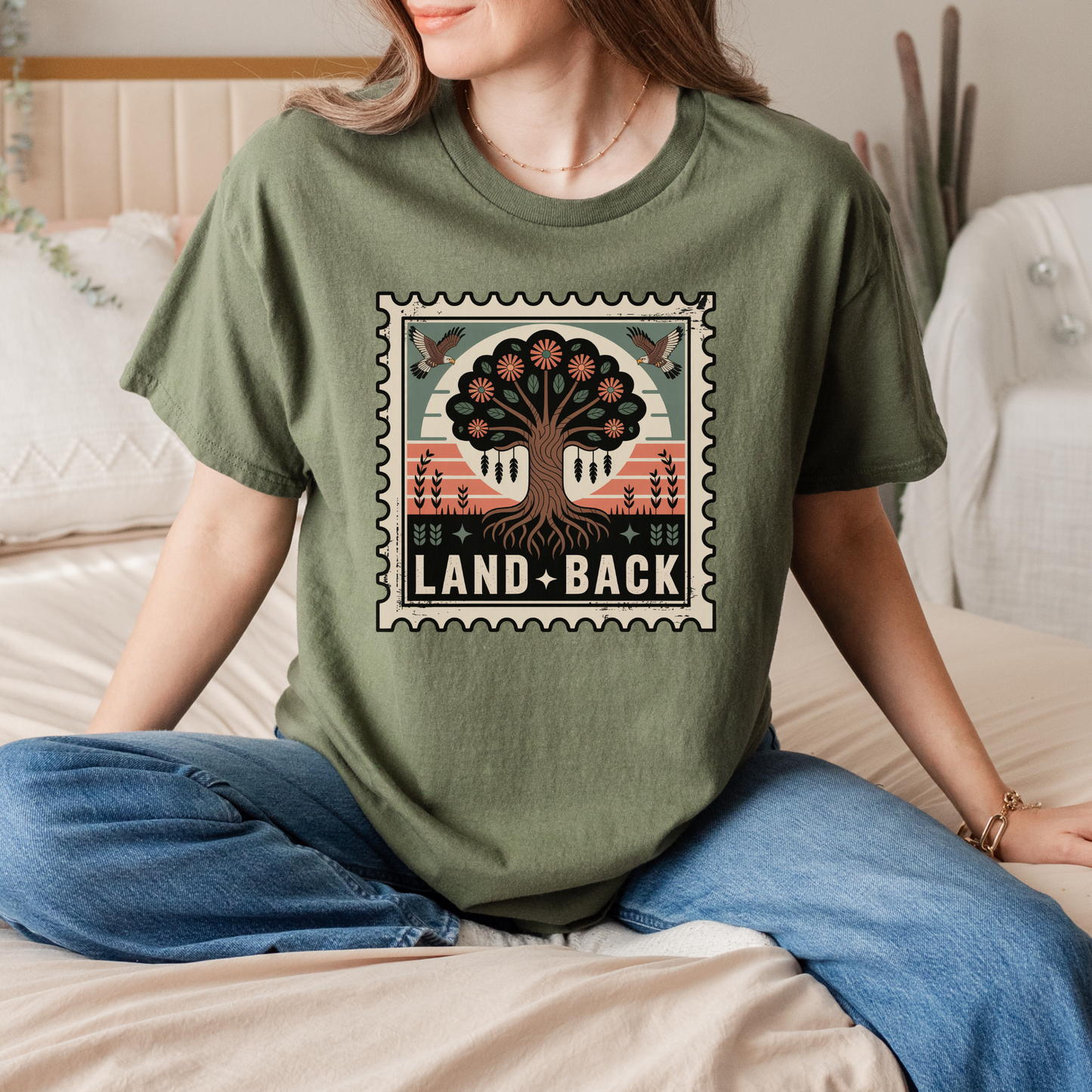 Person wearing a green t-shirt with a graphic design and text sitting on a bed. land back shirt, indigenous awareness shirt, nature shirt, tree graphic tee, minimalist shirt, advocacy apparel, eco conscious clothing, indigenous inspired apparel, social justice shirt, unisex t shirt, earthy aesthetic clothing, statement tee