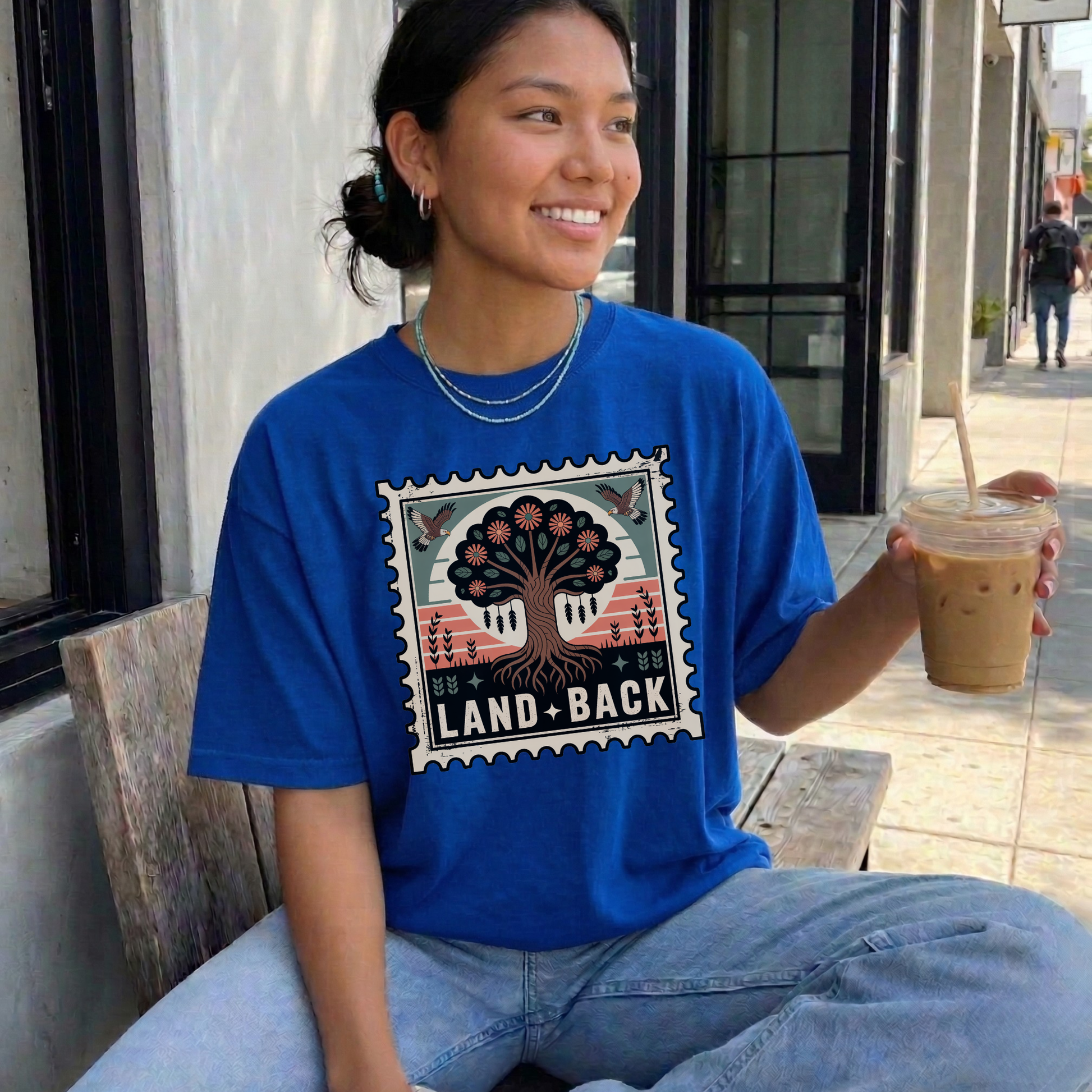 Person wearing a blue t-shirt with a graphic design and text, holding a drink outdoors. land back shirt, indigenous awareness shirt, nature shirt, tree graphic tee, minimalist shirt, advocacy apparel, eco conscious clothing, indigenous inspired apparel, social justice shirt, unisex t shirt, earthy aesthetic clothing, statement tee