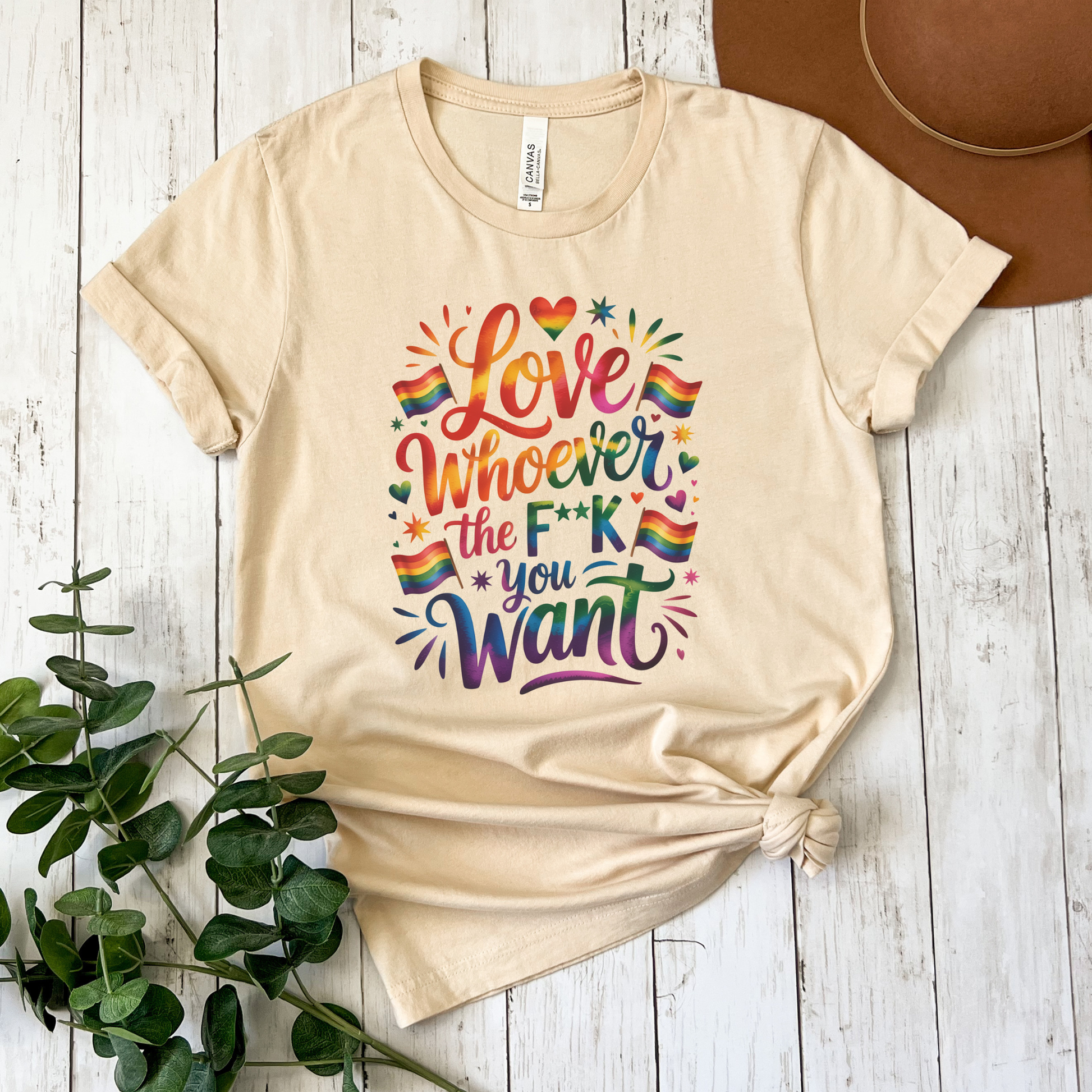 Beige t-shirt with colorful text and rainbow flags on a wooden surface with a plant and hat. love is love, pride shirt, equality apparel, inclusive clothing, feminist tee, graphic shirt, LGBTQ+ fashion, Indigenous brand, Toronto streetwear, statement tee, self-expression shirt