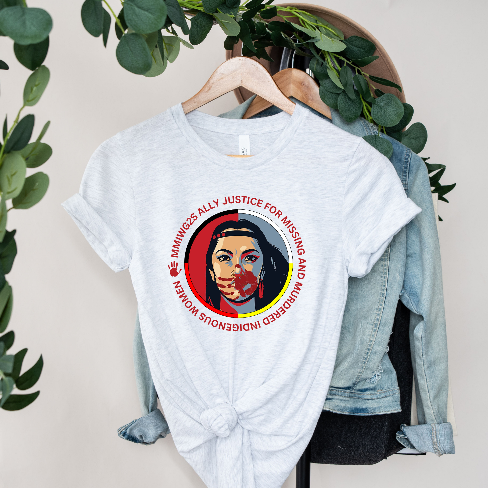 White t-shirt with a graphic design on a hanger with a denim jacket and greenery in the background. MMIWG2S shirt, Missing Murdered Indigenous Women tee, Indigenous ally shirt, MMIWG awareness apparel, justice for Indigenous women clothing, truth and reconciliation tee, National Day for Truth shirt, Indigenous activism clothing, 2SLGBTQQIA ally shirt, Canada missing women awareness, social justice tee, rally shirt Indigenous rights, solidarity shirt, awareness apparel Indigenous, activism t-shirt