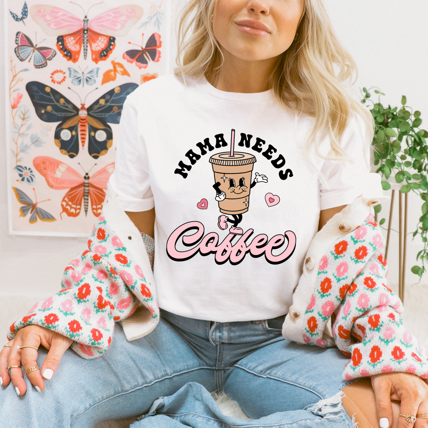 A woman wearing a white graphic t-shirt that reads 'MAMA NEEDS Coffee' with a cartoon coffee cup, styled with a pink floral cardigan and blue jeans, seated in front of a butterfly art print.