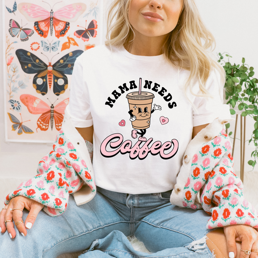 A woman wearing a white graphic t-shirt that reads 'MAMA NEEDS Coffee' with a cartoon coffee cup, styled with a pink floral cardigan and blue jeans, seated in front of a butterfly art print.