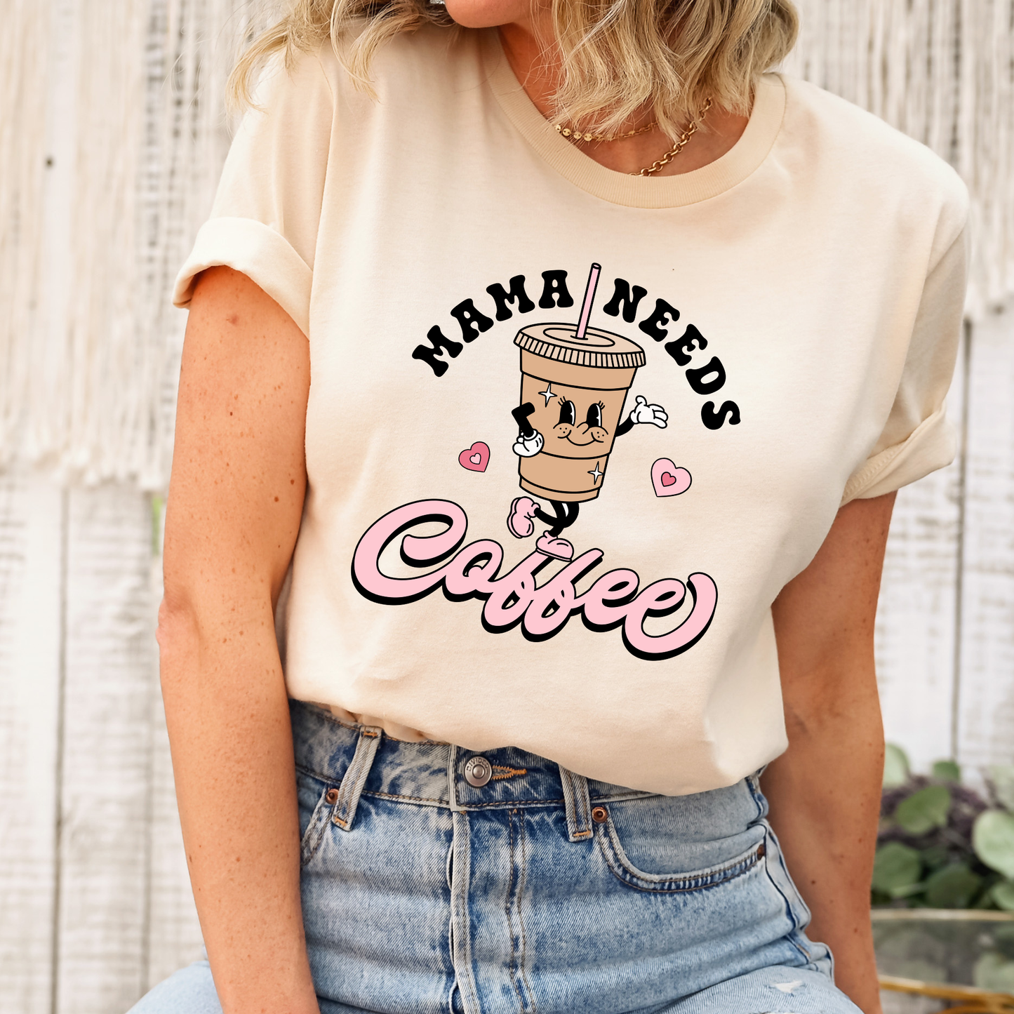 A woman wearing a cream-colored graphic t-shirt with rolled-up sleeves and light-wash blue jeans. The t-shirt features a cartoon coffee cup character with the text 'MAMA NEEDS Coffee' in black and pink lettering. mama needs coffee, coffee mom shirt, funny mom shirt, coffee lover gift, mom graphic tee, retro coffee shirt, caffeine shirt, mom life shirt, mothers day gift, cute coffee tee, aesthetic mom shirt, coffee aesthetic, sahdeh spruce