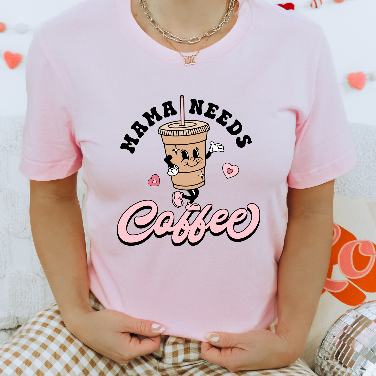 A person wearing a light pink short-sleeve graphic T-shirt with the text 'MAMA NEEDS Coffee' and a cartoon coffee cup character, seated indoors against a soft white background with decorative heart garlands.