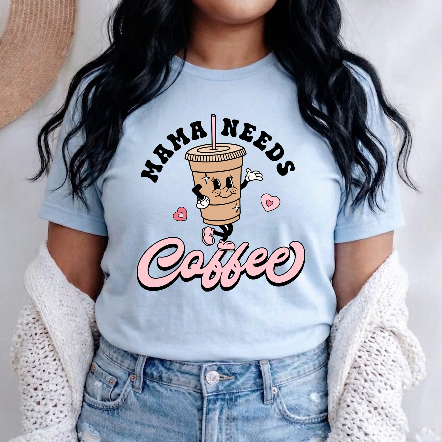 A woman wearing a light blue graphic t-shirt with a cartoon coffee cup and the text 'MAMA NEEDS Coffee' in pink and black. She is also wearing a white knit cardigan and light-wash blue jeans.
