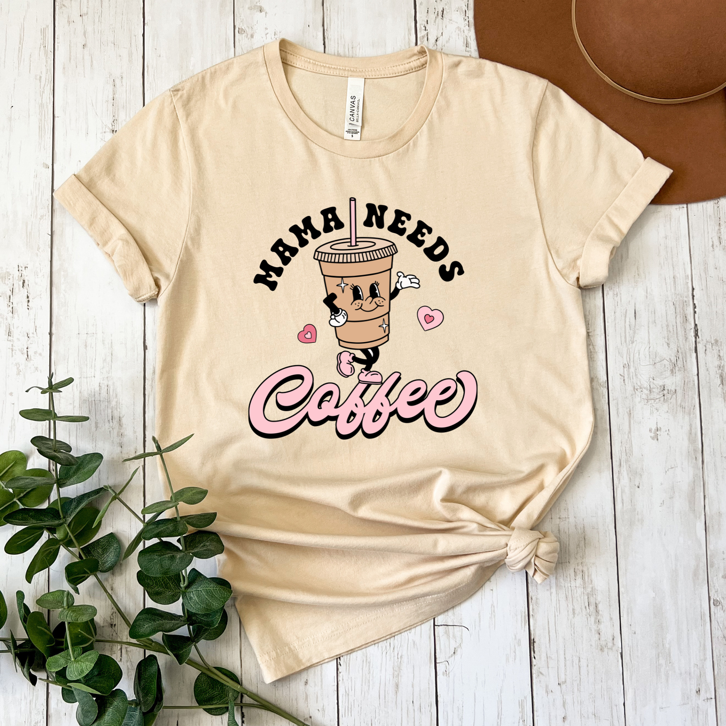 A beige graphic t-shirt featuring a cartoon coffee cup character and the text 'MAMA NEEDS Coffee' in black and pink, styled flat on a white wood plank background with a brown hat and green eucalyptus leaves.