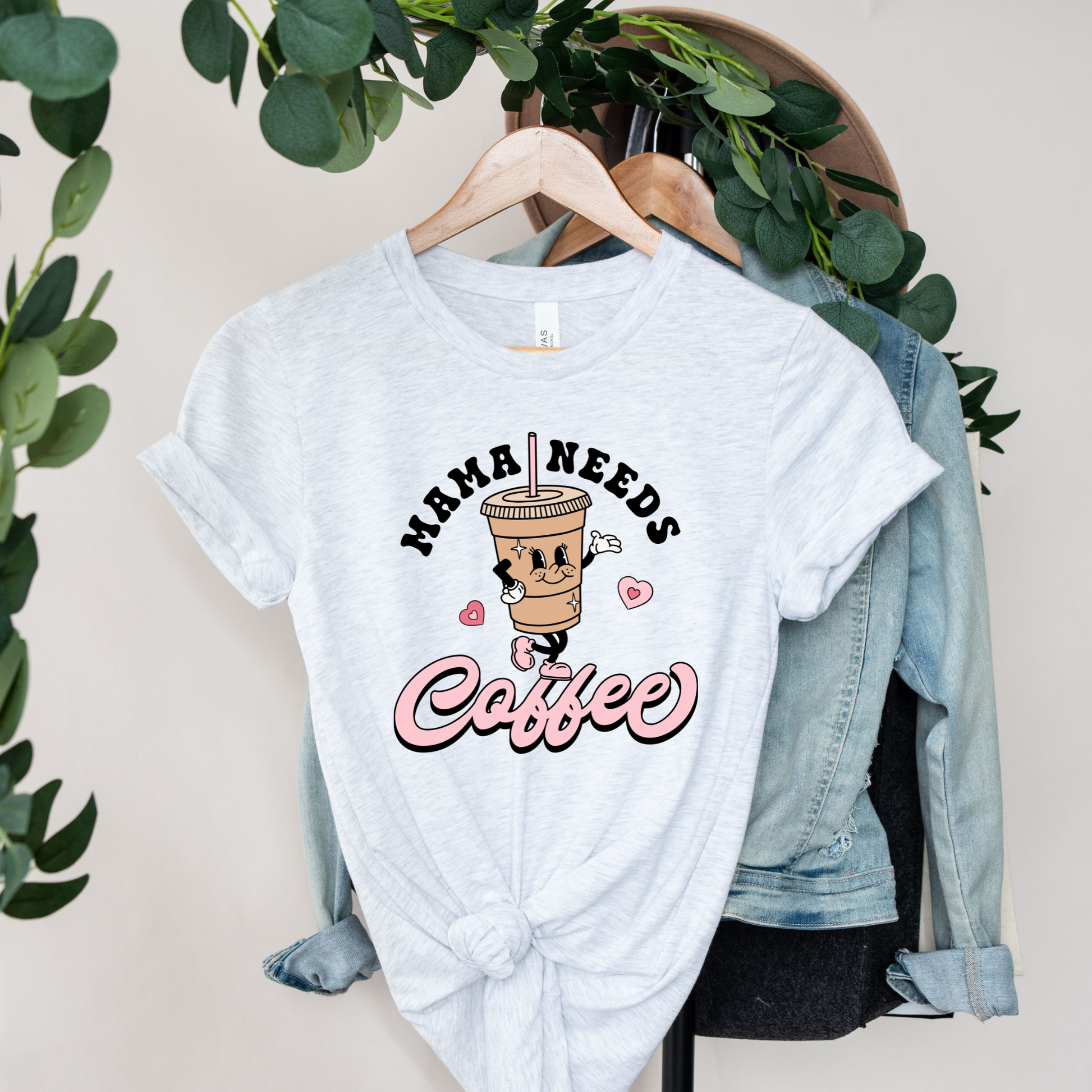 A light heather gray t-shirt with a 'HAMA NEEDS Coffee' graphic print, styled on a wooden hanger over a light blue denim jacket and black pants, with green foliage hanging in the background.
