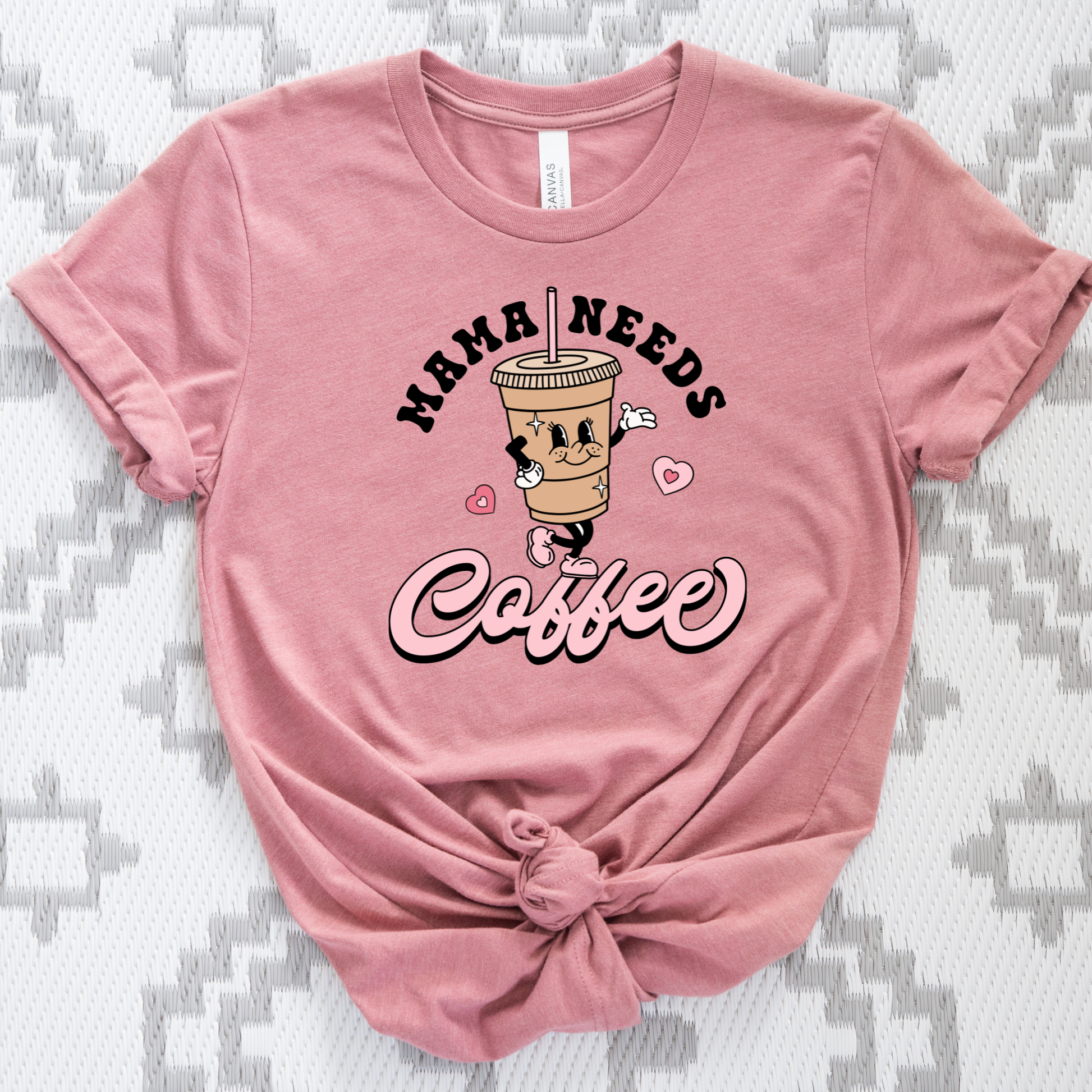A dusty pink short-sleeve t-shirt with a graphic of a cartoon coffee cup and the text 'MAMA NEEDS Coffee' in black and pink script. The shirt is styled with a knot at the bottom hem and is laid flat on a gray and white patterned fabric background.