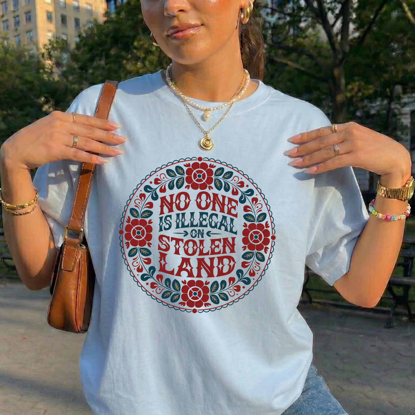 Person wearing a t-shirt with a floral design and text in an outdoor setting. No one is illegal on stolen land t-shirt, floral circle shirt, Indigenous floral tee, land back shirt, social justice t-shirt, stolen land circle design, activist floral apparel, Indigenous owned t-shirt, Sahdeh Spruce design, unisex activist tee, decolonization clothing, retro floral shirt, ethical fashion, protest graphic tee