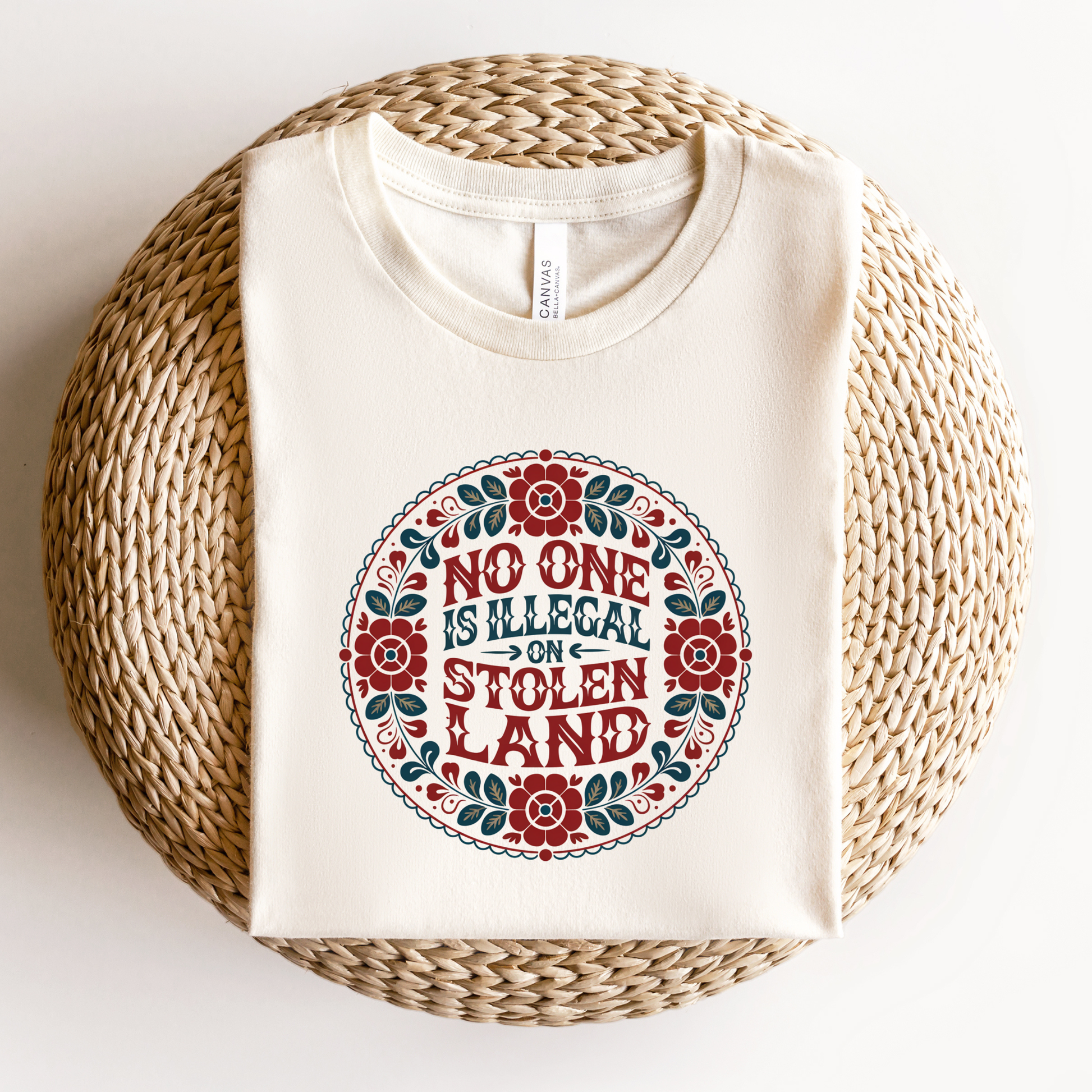 White t-shirt with 'No one is illegal on stolen land' design on a woven surface. No one is illegal on stolen land t-shirt, floral circle shirt, Indigenous floral tee, land back shirt, social justice t-shirt, stolen land circle design, activist floral apparel, Indigenous owned t-shirt, Sahdeh Spruce design, unisex activist tee, decolonization clothing, retro floral shirt, ethical fashion, protest graphic tee