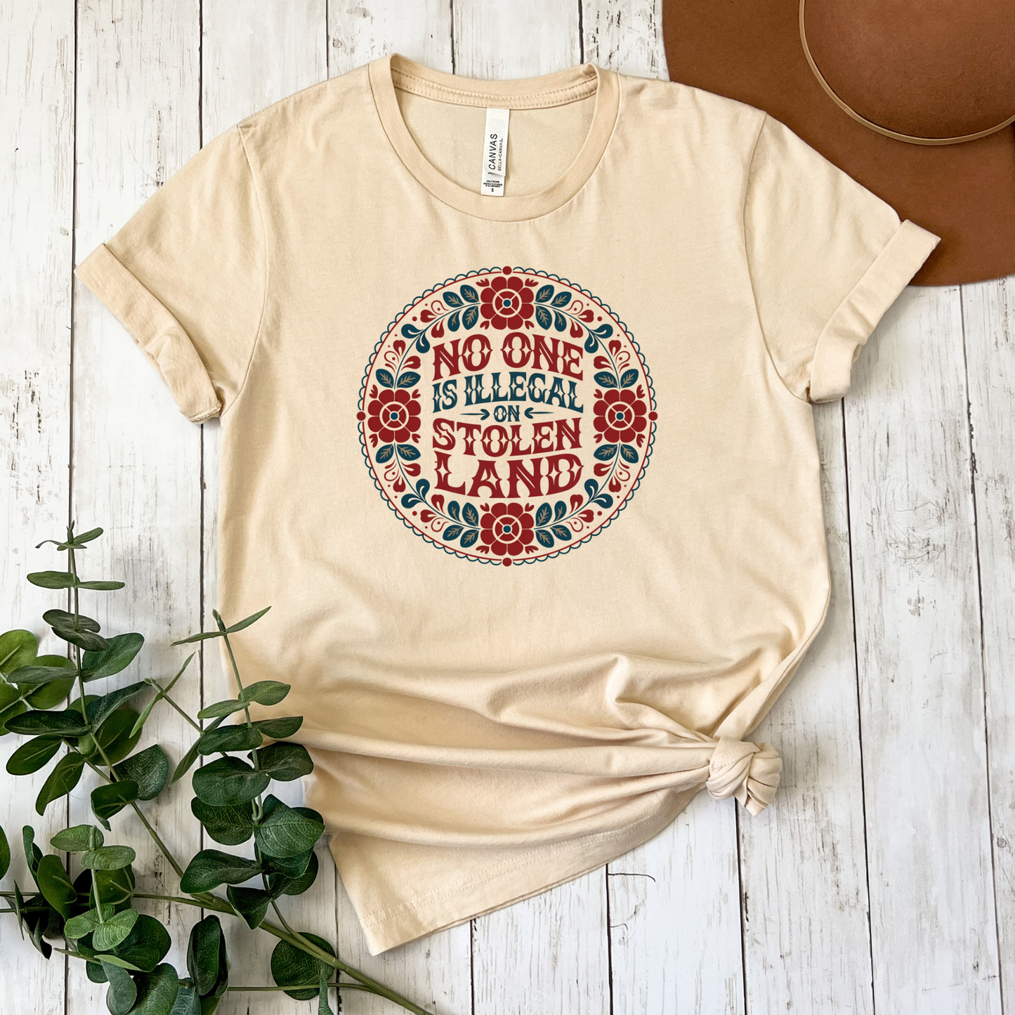 Beige t-shirt with a colorful graphic and text on a wooden surface with a plant and hat. No one is illegal on stolen land t-shirt, floral circle shirt, Indigenous floral tee, land back shirt, social justice t-shirt, stolen land circle design, activist floral apparel, Indigenous owned t-shirt, Sahdeh Spruce design, unisex activist tee, decolonization clothing, retro floral shirt, ethical fashion, protest graphic tee