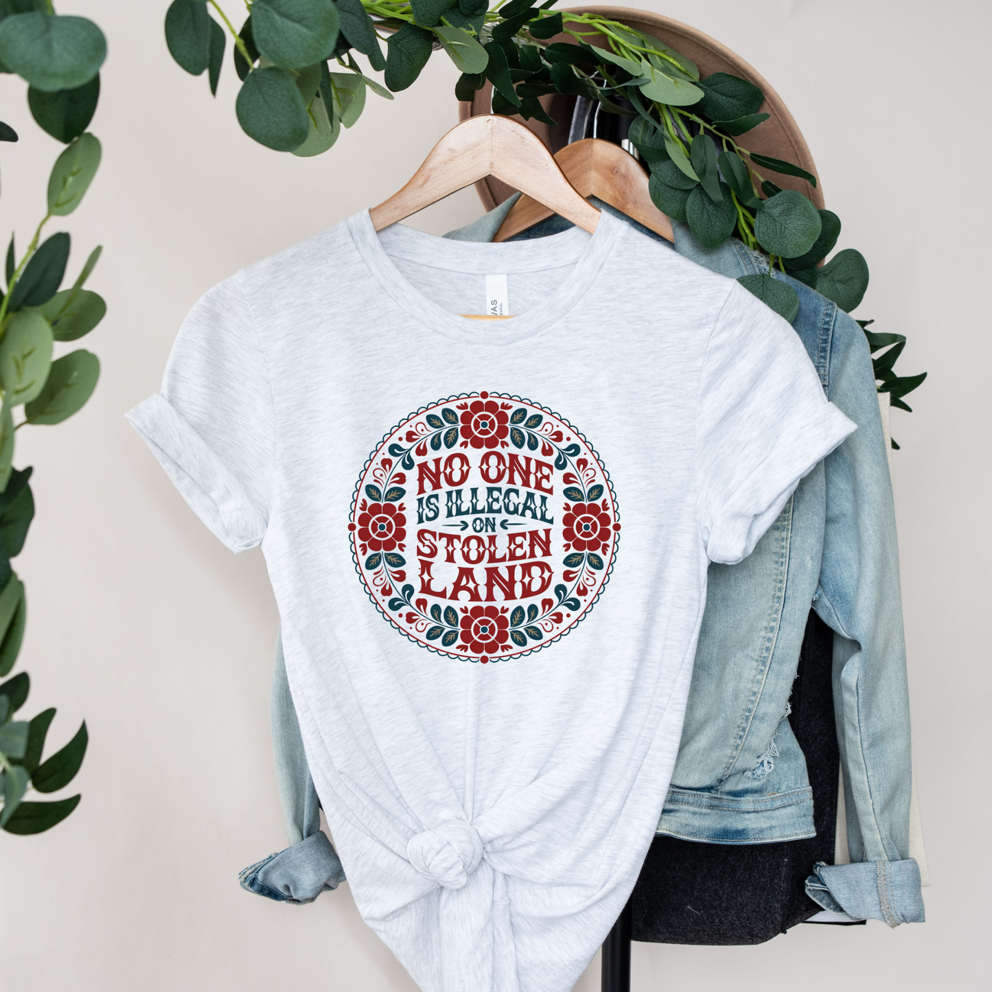 ash t-shirt with a floral design and text on a hanger with a denim jacket and greenery. No one is illegal on stolen land t-shirt, floral circle shirt, Indigenous floral tee, land back shirt, social justice t-shirt, stolen land circle design, activist floral apparel, Indigenous owned t-shirt, Sahdeh Spruce design, unisex activist tee, decolonization clothing, retro floral shirt, ethical fashion, protest graphic tee