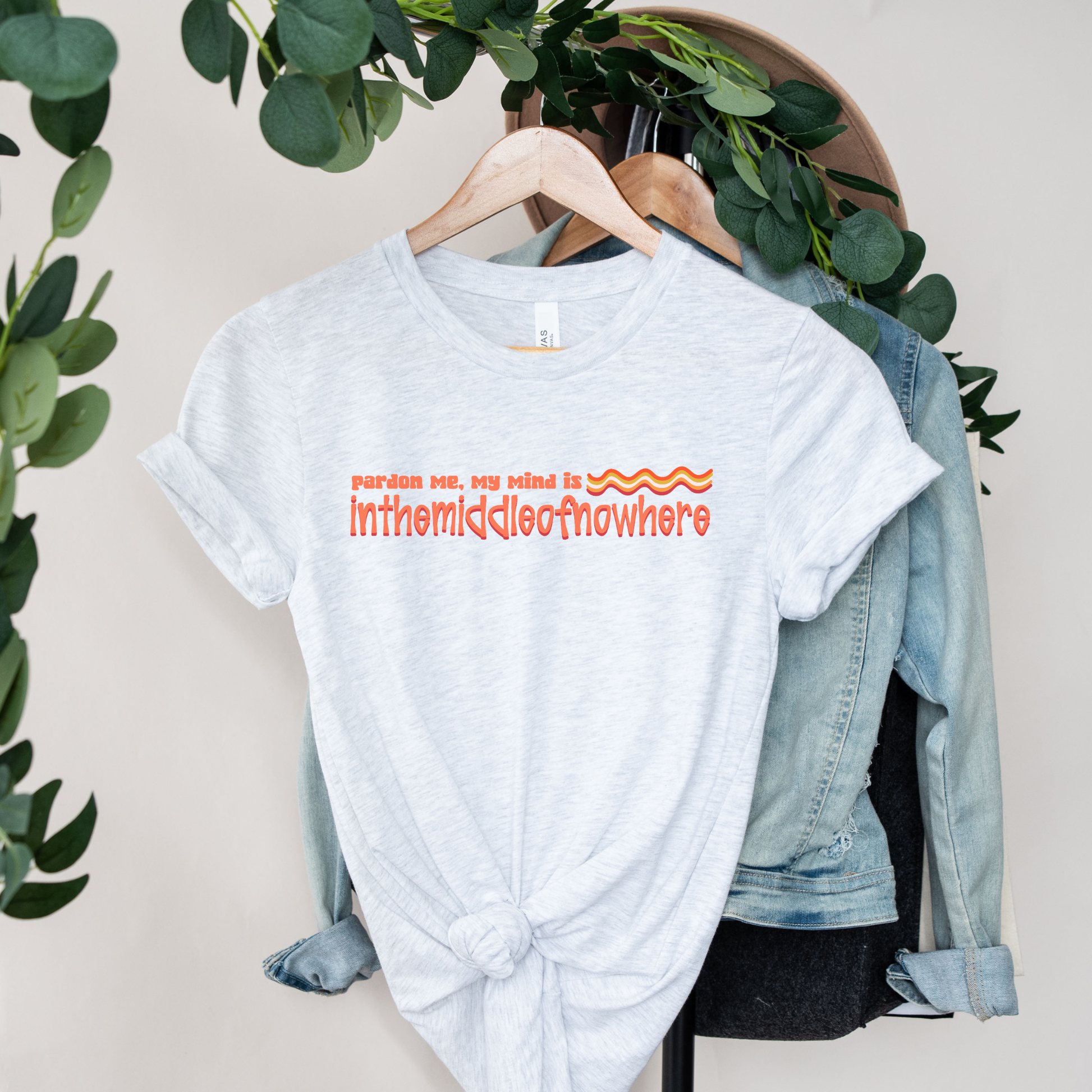 White t-shirt with orange text on a hanger with a denim jacket and greenery in the background funny quote shirt, sarcastic t shirt, unisex graphic tee, humor tee, funny saying shirt, casual quote shirt, statement tee, laid back shirt, daydreaming shirt, witty t shirt, gift idea shirt, funny unisex tee, everyday graphic tee, relaxed style shirt, humorous apparel