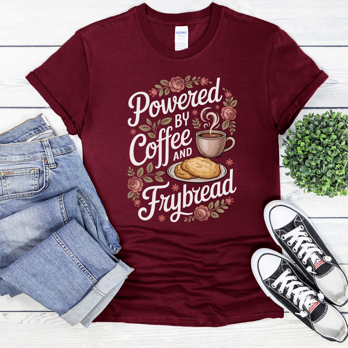 Maroon t-shirt with 'Powered by Coffee and Frybread' design on a white wooden surface with jeans and sneakers. coffee shirt, frybread shirt, indigenous shirt, funny indigenous shirt, coffee lover tee, cultural apparel, indigenous inspired clothing, casual graphic tee, funny t shirt, foodie shirt, native humor shirt, everyday wear tee, statement shirt, gift idea shirt