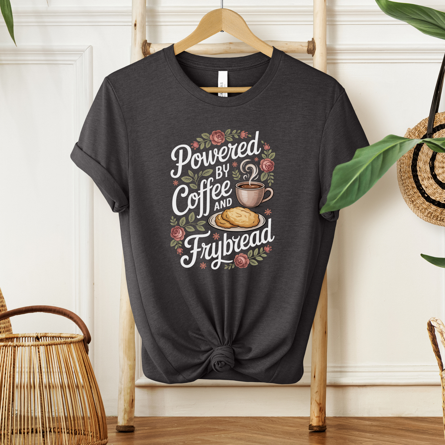 grey t-shirt with 'Powered by Coffee and Frybread' design on a wooden hanger. coffee shirt, frybread shirt, indigenous shirt, funny indigenous shirt, coffee lover tee, cultural apparel, indigenous inspired clothing, casual graphic tee, funny t shirt, foodie shirt, native humor shirt, everyday wear tee, statement shirt, gift idea shirt