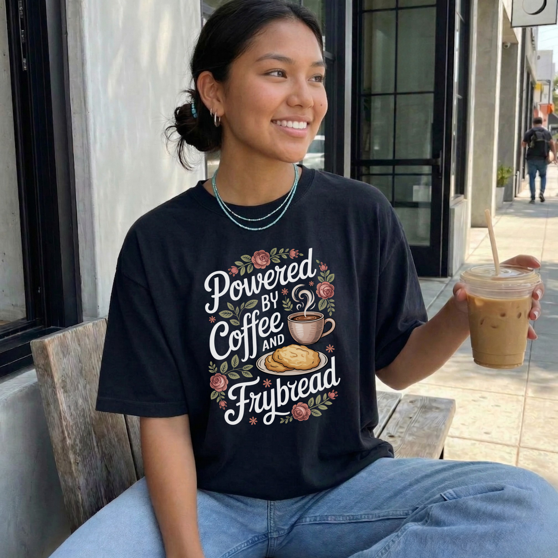 Woman wearing a black t-shirt with 'Powered by Coffee and Frybread' design, holding a drink outdoors.  coffee shirt, frybread shirt, indigenous shirt, funny indigenous shirt, coffee lover tee, cultural apparel, indigenous inspired clothing, casual graphic tee, funny t shirt, foodie shirt, native humor shirt, everyday wear tee, statement shirt, gift idea shirt