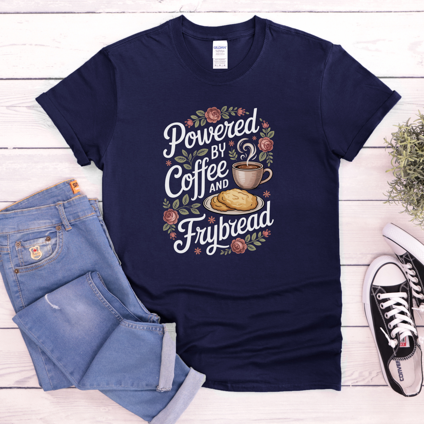 Navy blue t-shirt with 'Powered by Coffee and Frybread' design on a wooden surface with jeans and sneakers.coffee shirt, frybread shirt, indigenous shirt, funny indigenous shirt, coffee lover tee, cultural apparel, indigenous inspired clothing, casual graphic tee, funny t shirt, foodie shirt, native humor shirt, everyday wear tee, statement shirt, gift idea shirt
