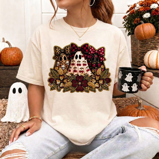 A woman wearing a cream-colored t-shirt with a Halloween-themed graphic print featuring three ghosts, pumpkins, and autumn leaves. She is holding a black mug with white ghost illustrations, set in a cozy fall-themed environment with pumpkins and a small ghost decoration.