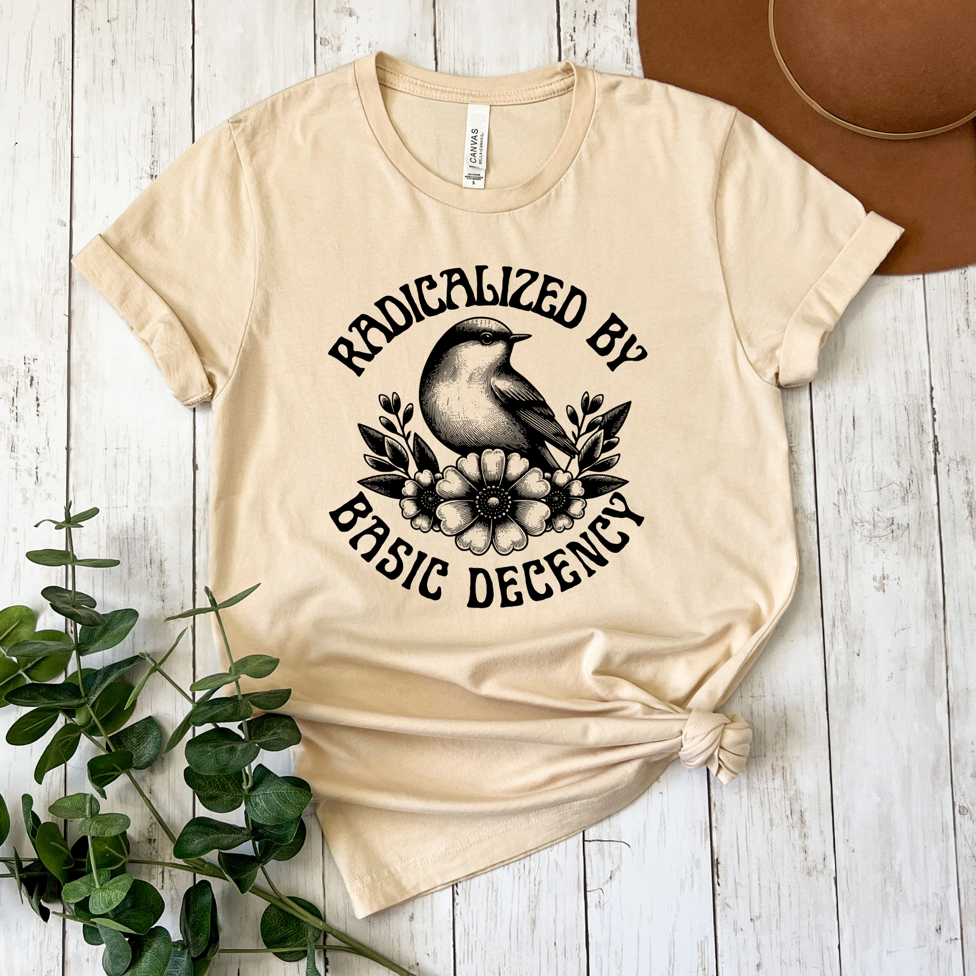 Beige t-shirt with graphic design and text on a wooden surface with a plant and hat. radicalized by basic decency shirt, progressive tee, activist shirt, social justice clothing, political statement tee, hypocrisy shirt, progressive apparel, rally shirt, protest tee, witty activist shirt, equality shirt, ally shirt, conversation starter tee, bold statement clothing, unisex political shirt