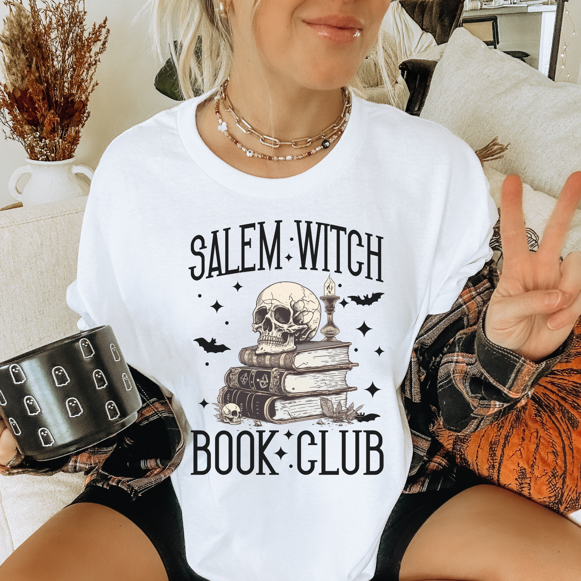 A woman wearing a white graphic t-shirt with a black 'Salem Witch Book Club' design featuring a skull, books, and candles. She is holding a black mug with tombstone patterns and wearing a plaid shirt underneath.