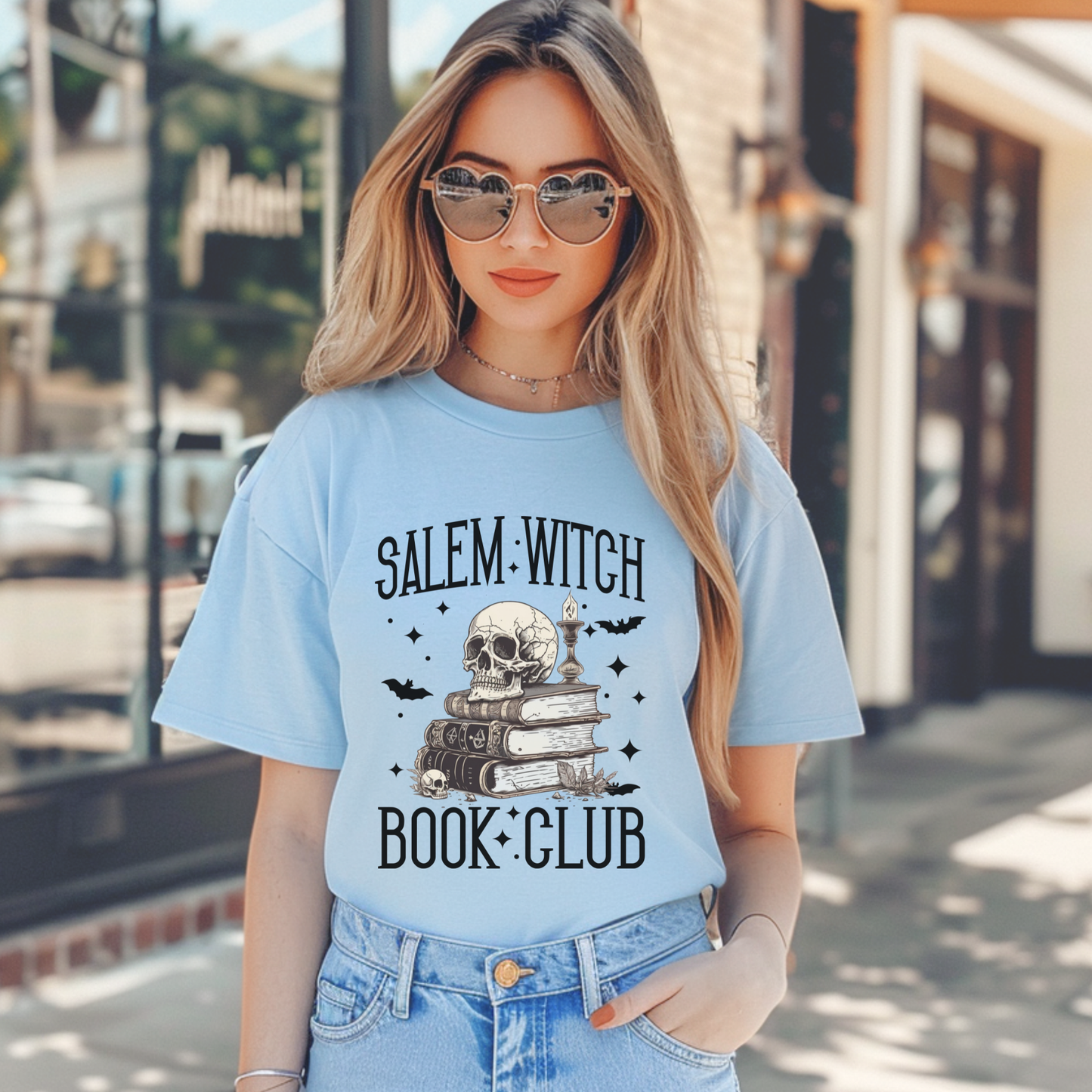 A woman wearing a light blue graphic t-shirt with a 'Salem Witch Book Club' design featuring a skull, books, and bats, paired with blue jeans in an outdoor street setting.