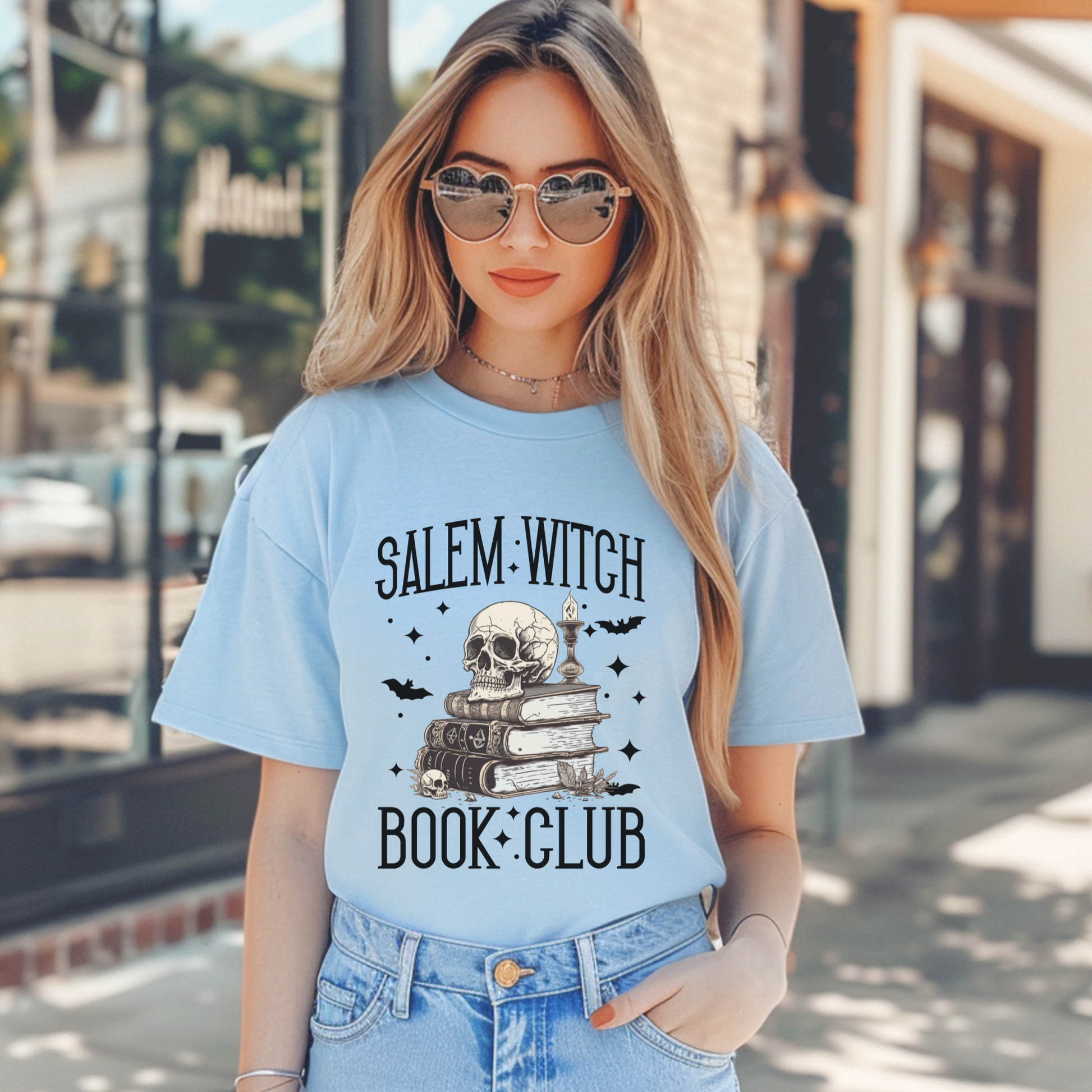 A woman wearing a light blue graphic t-shirt with a 'Salem Witch Book Club' design featuring a skull, books, and bats, paired with blue jeans in an outdoor street setting.