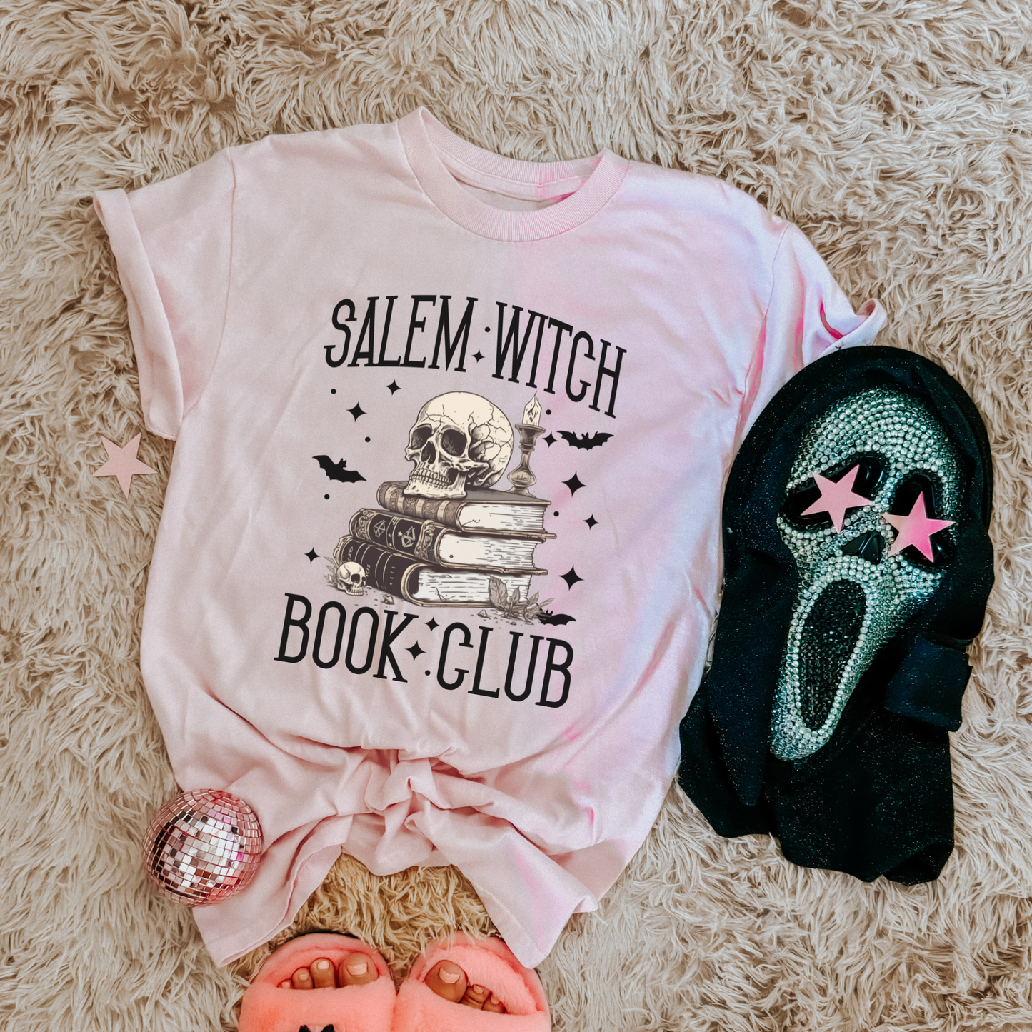 A styled flat-lay of a light pink graphic T-shirt with 'SALEM WITCH BOOK CLUB' text and a skull-and-books design, arranged on a shag rug with a black Ghostface mask, pink slide sandals, and a pink sequined accessory.
