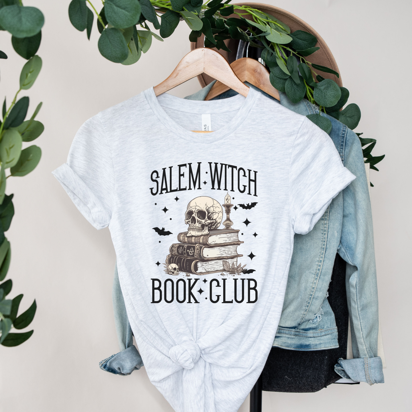 A light heather gray graphic T-shirt hanging on a wooden hanger, styled with a knotted hem and layered over a light blue denim jacket. The shirt features a graphic of a skull on a stack of books with the text 'SALEM WITCH BOOK CLUB' and small black bat silhouettes.