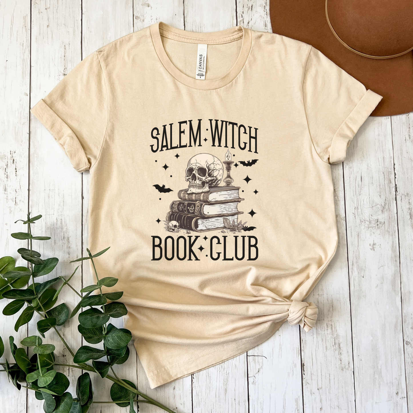 A beige graphic t-shirt featuring a 'Salem Witch Book Club' design with a skull, books, and bats, styled flat-lay on a white wood background with a brown hat and green leaves.