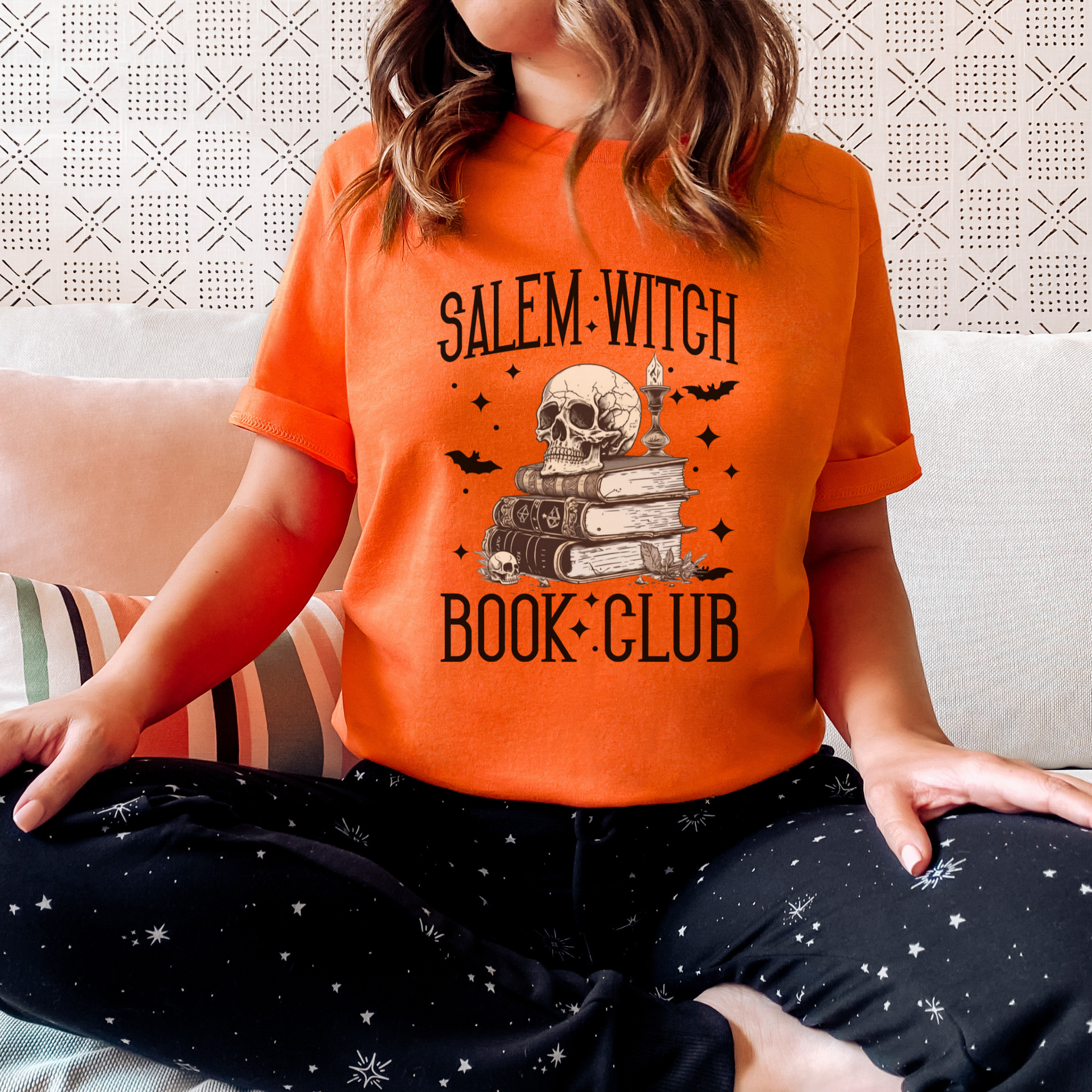 A woman wearing a bright orange t-shirt with a 'Salem Witch Book Club' graphic featuring a skull, books, and candles, paired with black starry pajama pants, sitting on a couch.