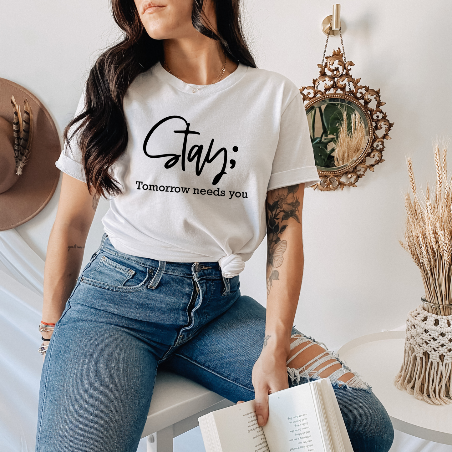 A woman wearing a white graphic t-shirt with the text 'Stay; Tomorrow needs you' in black script and sans-serif fonts, paired with blue jeans, sitting in a cozy, styled room setting.