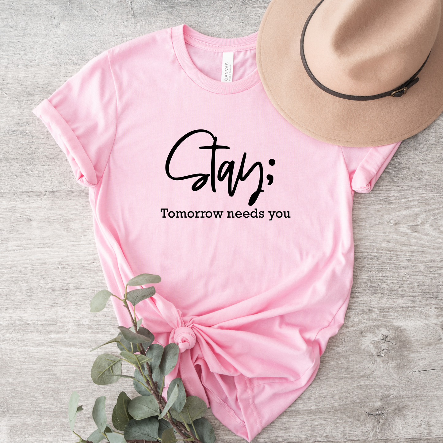 A pink short-sleeve graphic T-shirt laid flat on a light wood surface, styled with a tan hat and greenery. The shirt has black text reading 'Stay;' and 'Tomorrow needs you.