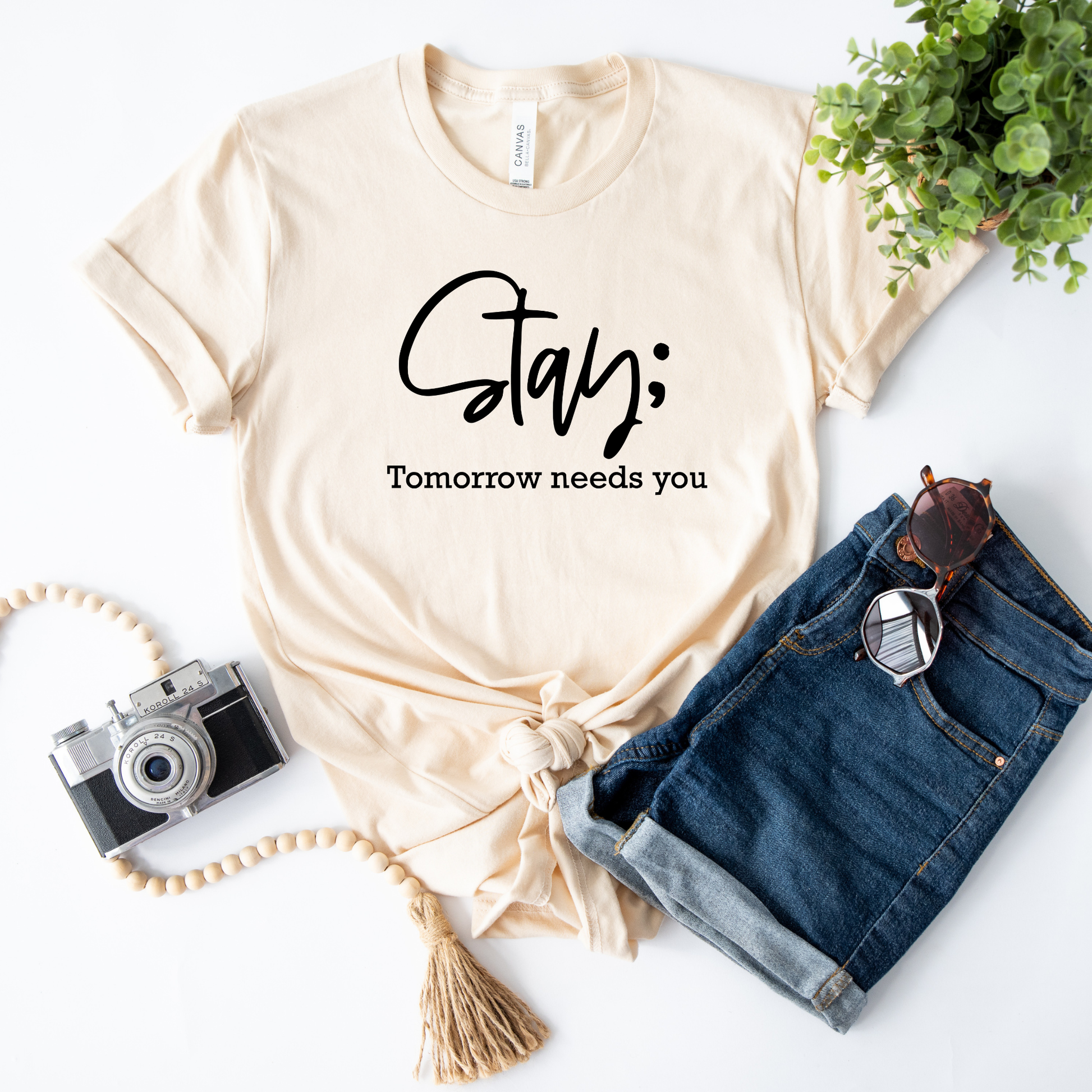 A cream-colored t-shirt with the text 'Stay; Tomorrow needs you' printed in black, styled in a flat-lay arrangement with blue denim shorts, sunglasses, a camera, and a tassel necklace on a white background.