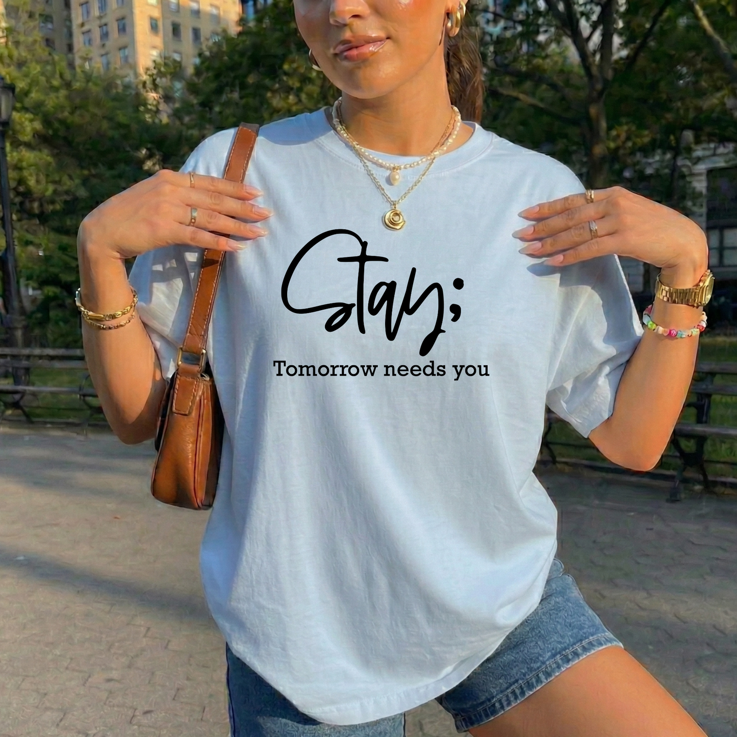 A woman wearing a light blue graphic t-shirt with the text 'Stay; Tomorrow needs you' printed in black. She is posing outdoors in a park setting with trees and benches in the background, wearing denim shorts and carrying a brown leather shoulder bag.