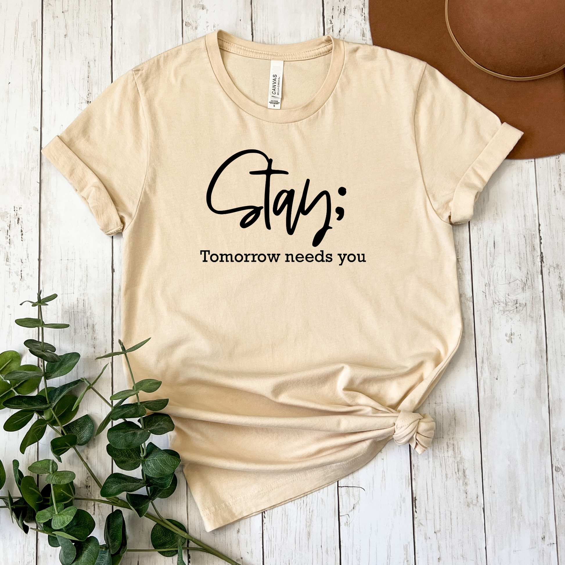 A beige graphic t-shirt with rolled sleeves, featuring the text 'Stay;' in black cursive and 'Tomorrow needs you' in black serif font. The t-shirt is styled in a flat-lay arrangement on a white wooden background, with a brown hat in the top right corner and green eucalyptus leaves in the bottom left corner.